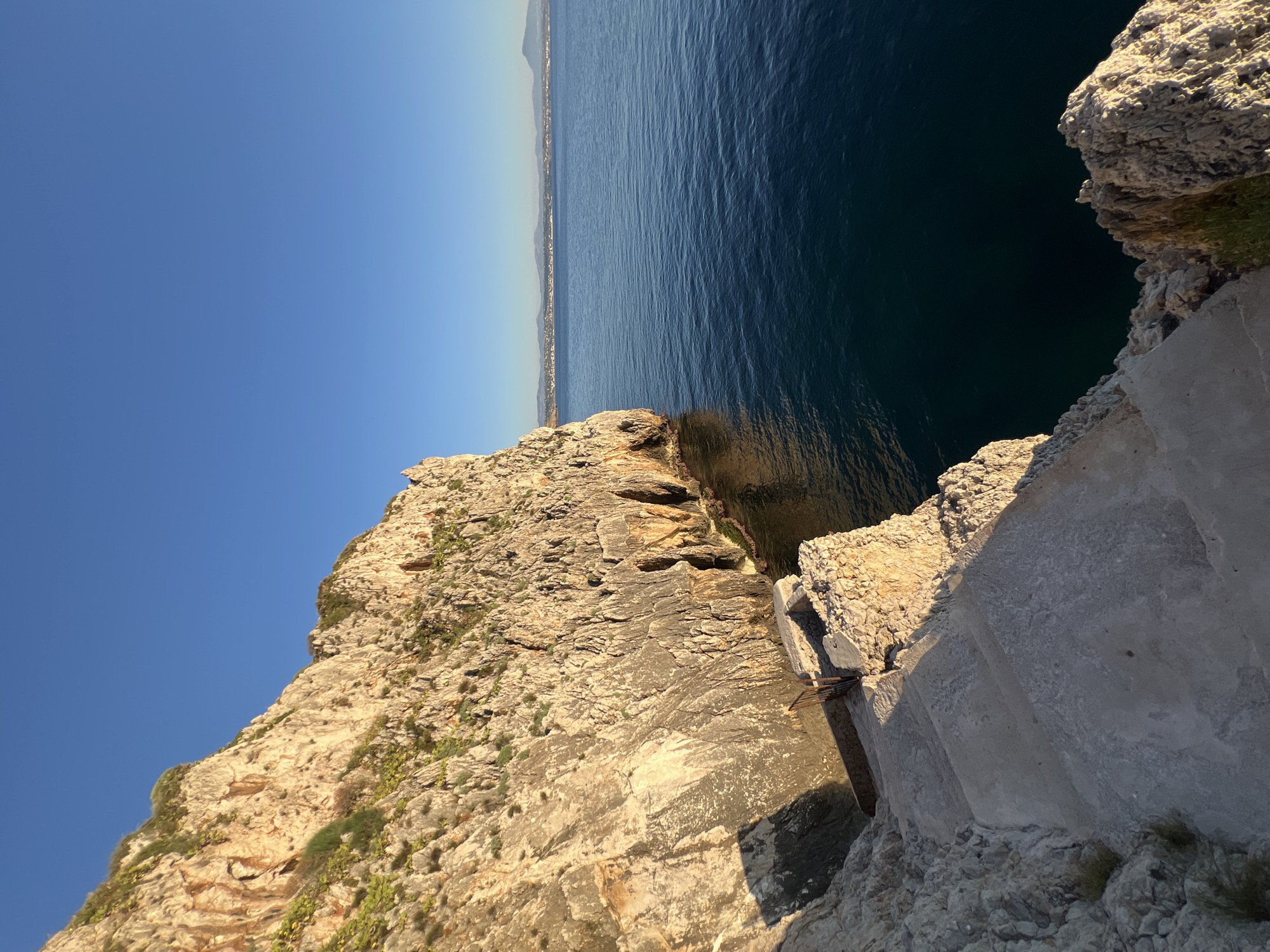 The image depicts a rocky cliff face overlooking a calm, dark blue sea. A stone staircase descends along the cliff's edge, with a rusty metal railing visible at the top. The sun casts long shadows, indicating it is either early morning or late afternoon. The cliff is sparsely covered with green vegetation. In the distance, a coastline with buildings and mountains can be seen under a clear blue sky. The scene is located in Terrasini, Italy. There are no people or animals visible in the image.