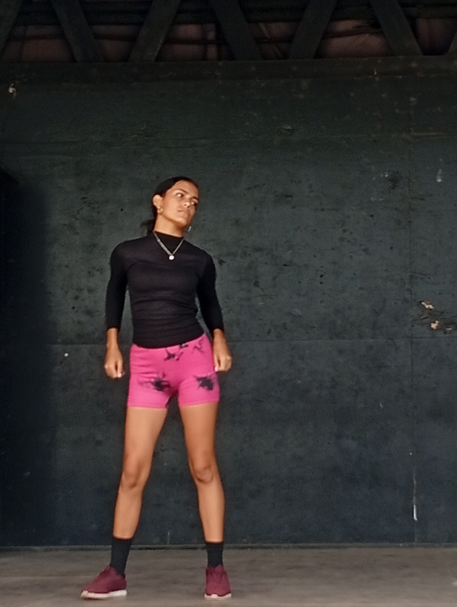 A woman stands in what appears to be an indoor or covered structure with a dark background. She is wearing a black long-sleeved top, pink shorts, black socks, and dark athletic shoes. She is looking off to her right. The background is a dark wall with some structural elements visible at the top, possibly indicating a roof or ceiling. The floor appears to be concrete or another hard surface. The location is likely Playa El Agua, Venezuela.