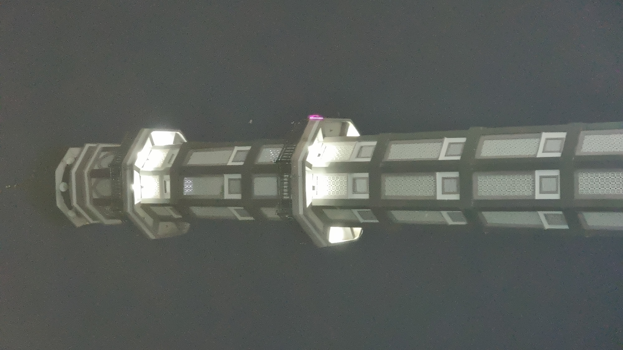 The image is a low-angle shot of a brightly lit minaret against a dark, hazy sky. The minaret is octagonal in shape and features multiple tiers with decorative window panels. Lights are visible emanating from within the structure, illuminating the facade and casting a glow on the surrounding mist. There are no people or other visible subjects in the image. The overall atmosphere is subdued due to the overcast conditions and the monochromatic color palette dominated by grays and white. The location is identified as Keudekruenggeukueh, Indonesia.