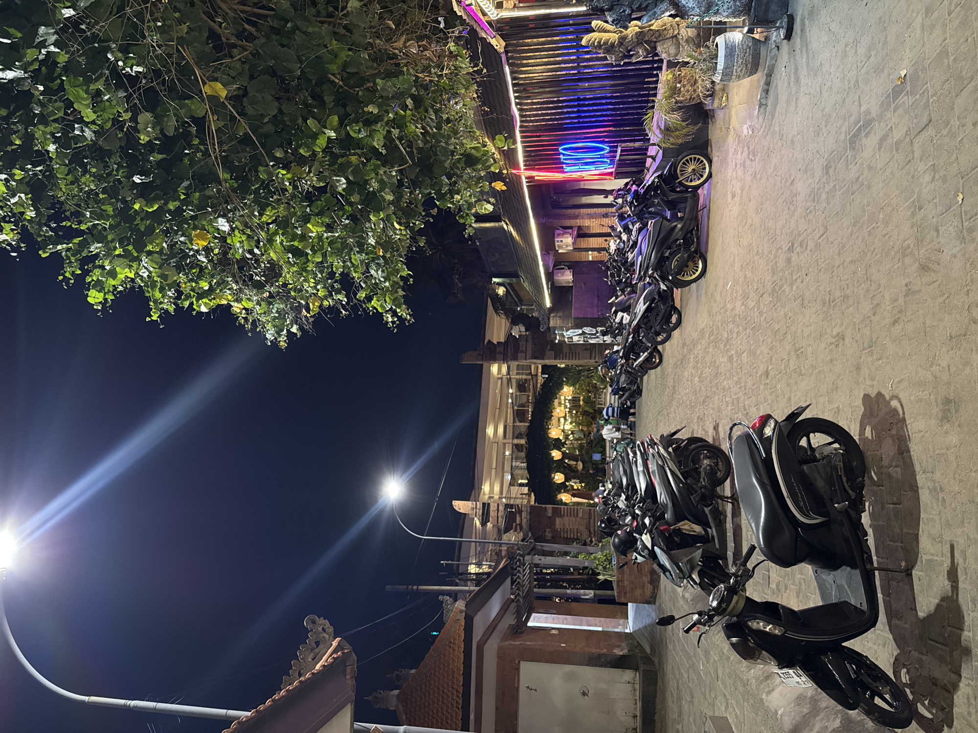 This is a night photograph of a street in Kuta, Indonesia, characterized by a row of parked scooters. The street is paved with interlocking bricks, and a tall street lamp illuminates the scene with a bright, cool light, casting long shadows. To the left, a traditional building with an ornate roofline, reminiscent of Balinese architecture, is visible. Further down the street, a building with a wooden facade is lit by neon signs, one of which spells out "NEO" in pink and blue. A large, leafy tree hangs over the scene from the right. In the background, several people are gathered, some appearing to be staff or patrons of the establishment with the neon sign. The overall atmosphere is lively and urban, typical of a night in a tourist area.