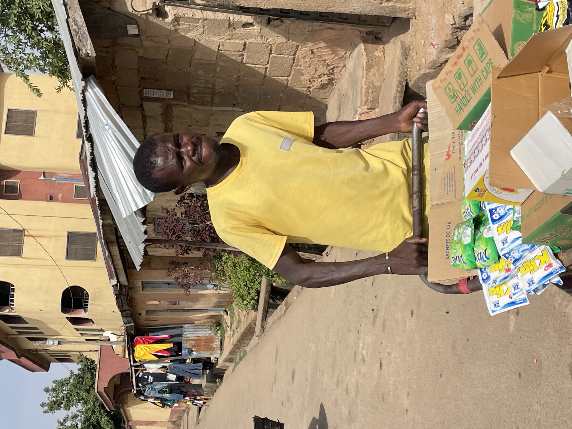 A man wearing a yellow t-shirt is pulling a cart filled with boxes. The cart is loaded with various items, including boxes with the words "HANDLE WITH CAPE", "SACHETS DE 12g" and "STORE IN A DRY". He is standing on a paved street with buildings and clotheslines visible in the background. The location appears to be a city street in Unknown, Unknown.