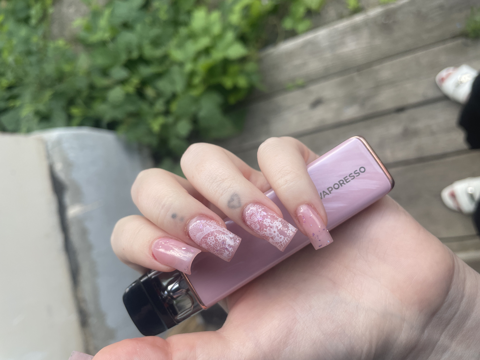 The image shows a close-up of a person's hand with long, pink, square-shaped artificial nails, decorated with white lace patterns and glitter. The hand is holding a pink Vaporesso vape device. The background is slightly blurred, showing greenery, wooden planks, and what appears to be a concrete structure. The lighting suggests it's daytime, possibly outdoors. There are no clear indicators of weather or specific emotions. Visible text on the vape device reads "VAPORESSO". The scene suggests a casual moment, potentially outdoors, given the background elements.