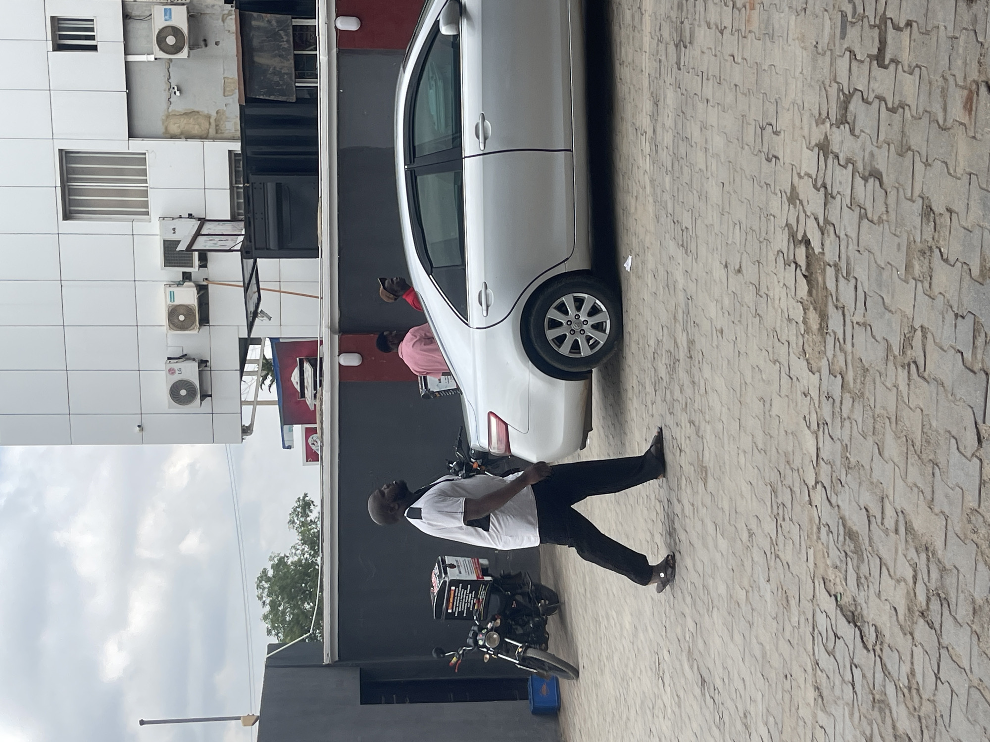 The image shows a daytime scene in Galadimawa Village, Nigeria. A white car is parked on a paved surface made of interlocking bricks. A man wearing a white shirt and black pants is walking away from the car. A motorcycle is parked behind the car. Two people stand near the car. The background includes a tall, light-colored building with multiple air conditioning units mounted on the wall. There is also a dark grey wall and some signage visible. The sky is overcast.