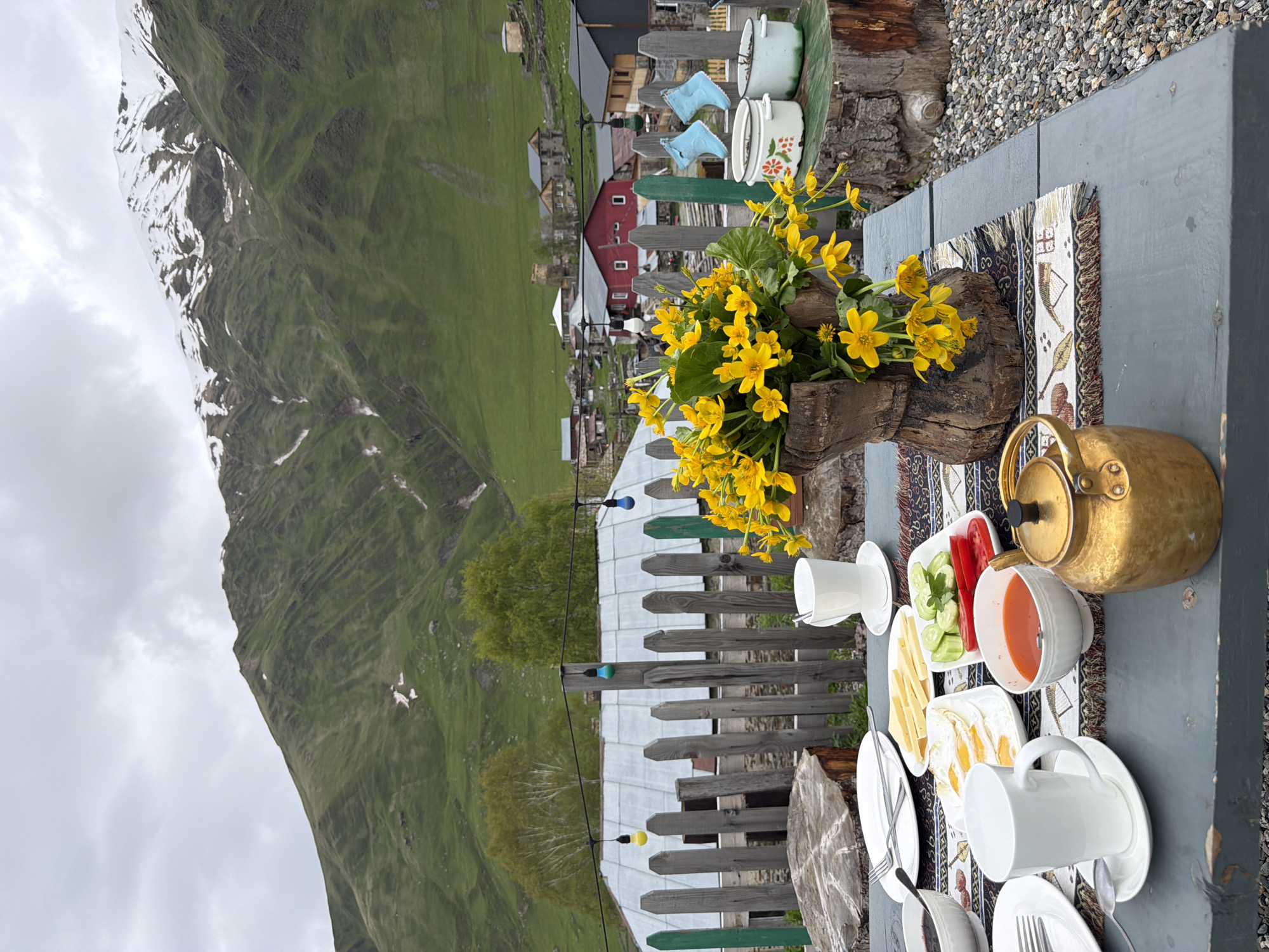 The image depicts an outdoor breakfast table set in the village of Ushguli, Georgia. A grey wooden table holds plates of food including fried eggs, sliced cheese, cucumbers, tomatoes, and a bowl of red soup. Two white mugs and a golden teapot are also present. A vase filled with yellow flowers sits in the foreground. In the background, a wooden fence separates the table from a village with traditional houses and a backdrop of green, snow-capped mountains under a cloudy sky.