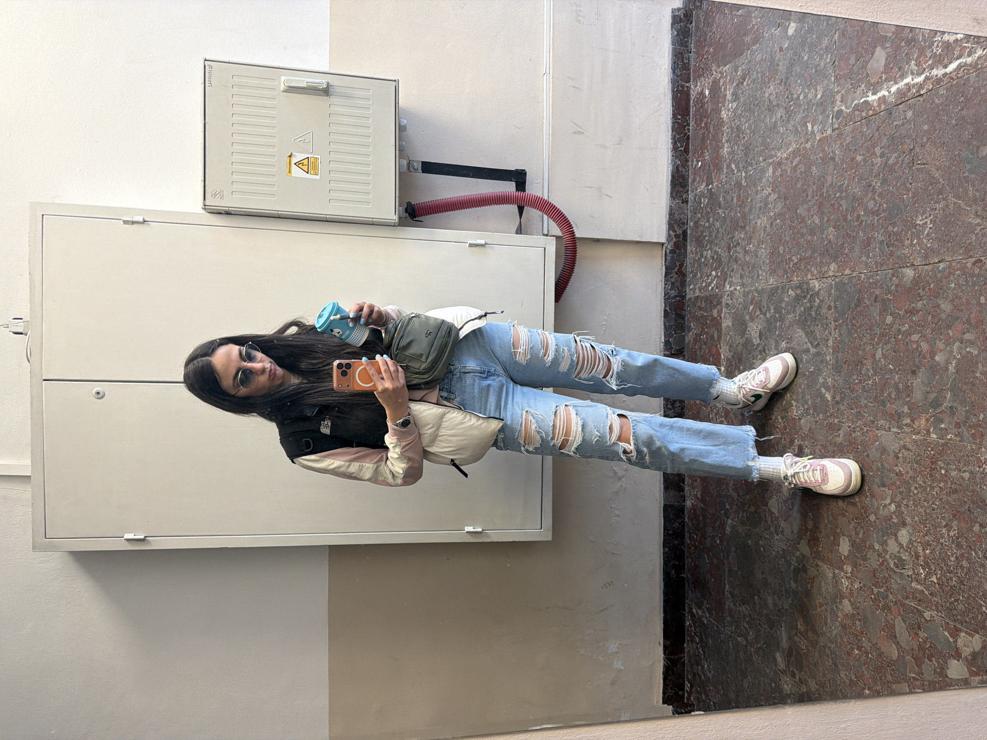 A young woman is taking a selfie in a hallway. She's holding a phone in one hand and a blue coffee cup in the other. She's wearing ripped jeans, sneakers, a puffer jacket, and sunglasses. The hallway has a white wall with electrical boxes and a marble floor. This is likely a public building. The location is most likely Sofia, Bulgaria.