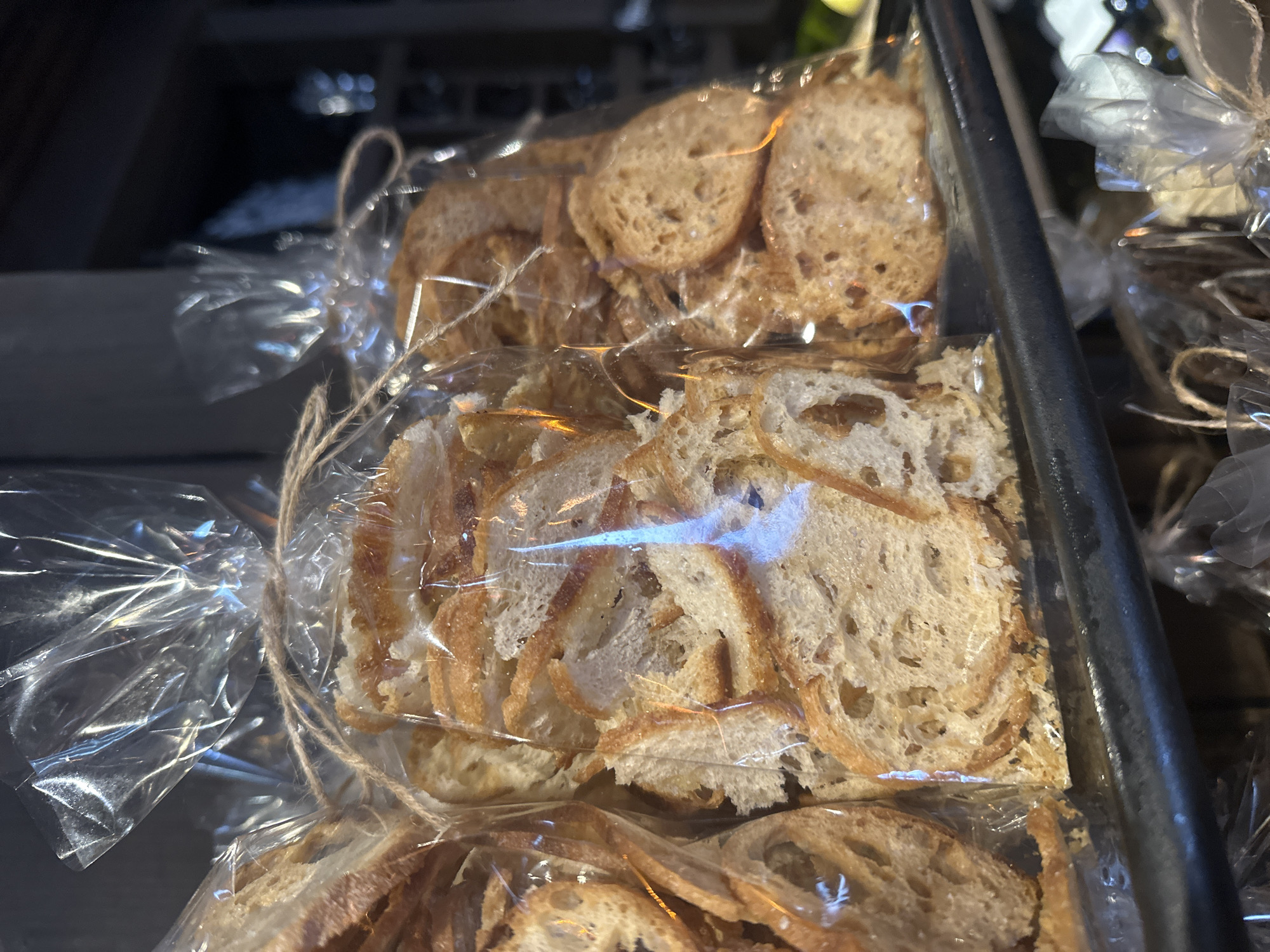 The image depicts several clear plastic bags filled with toasted bread slices, likely crostini or similar. Each bag is tied at the top with a simple string or twine. The bread slices are of varying colors, ranging from light tan to darker brown, suggesting different degrees of toasting. The bags are displayed behind a metal bar, likely in a store or bakery setting. Other similar bags are visible in the background. The image seems to be taken indoors, possibly in Новоглаголево, Russia.