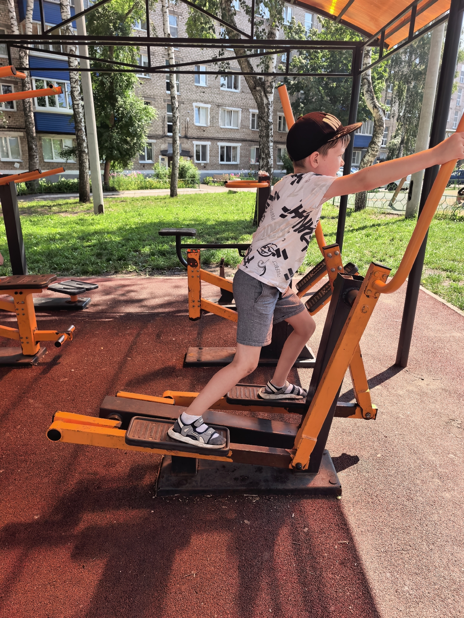 A young boy, wearing a black baseball cap, a white t-shirt with black graphics, grey shorts, and sandals, is using an orange and black outdoor elliptical machine. He is positioned on the right side of the frame, his body angled towards the right. His right arm is extended forward, gripping a handle. His left leg is forward on the machine's pedal, and his right leg is back. The elliptical machine is situated on a red rubberized surface, commonly found in children's playgrounds or outdoor fitness areas. In the background, green grass and trees are visible, with a multi-story brick building behind them, featuring white-framed windows and blue accents. The scene is illuminated by bright sunlight, casting distinct shadows on the ground. This scene is located in a courtyard in Sterlitamak, Russia.