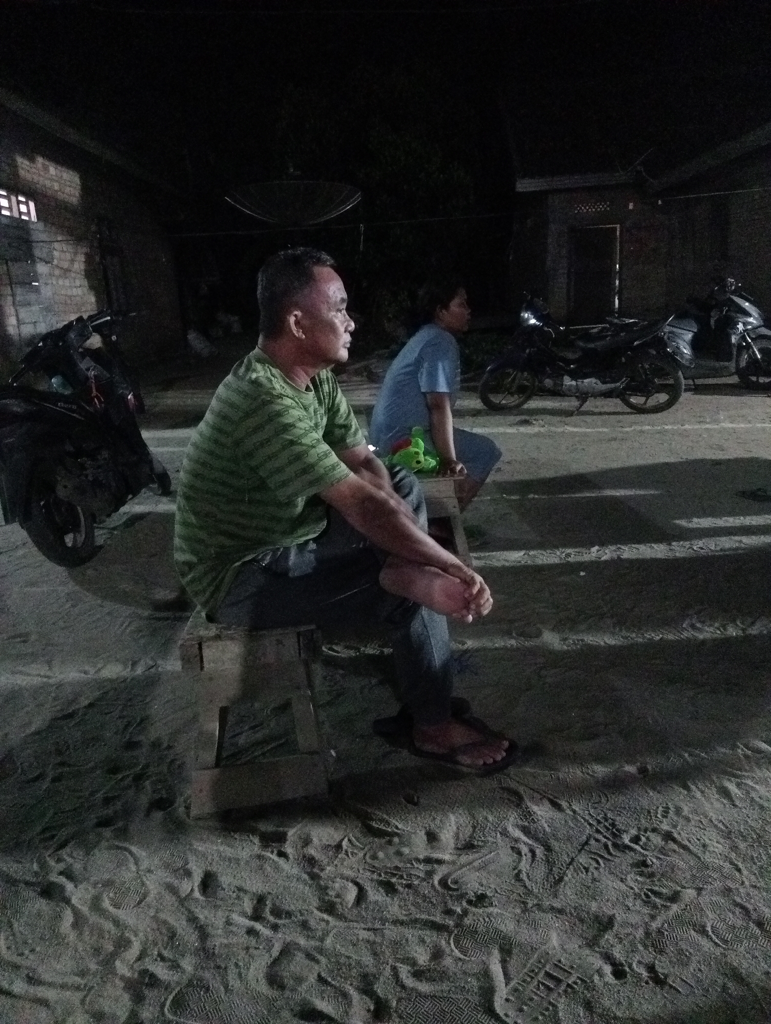 The image depicts an outdoor scene at night, likely in Rasuan Baru, Indonesia. In the foreground, a man wearing a green striped t-shirt and dark pants sits on a wooden bench. His body is angled towards the right, with his head turned to the side, suggesting he is looking at something out of frame. His hands are clasped in his lap, and his feet are resting on the ground. A second individual, partially visible, is seated on the same bench to the man's right. This person is wearing a light blue shirt and dark shorts, with a green stuffed toy placed beside them on the bench.
The environment appears to be sandy or dusty, with numerous footprints visible on the ground. Behind the seated individuals, several motorcycles are parked. The background includes a dimly lit building with a satellite dish mounted on it. The overall lighting suggests artificial illumination from an unseen source, casting shadows on the ground. The scene conveys a sense of quiet waiting or observation.