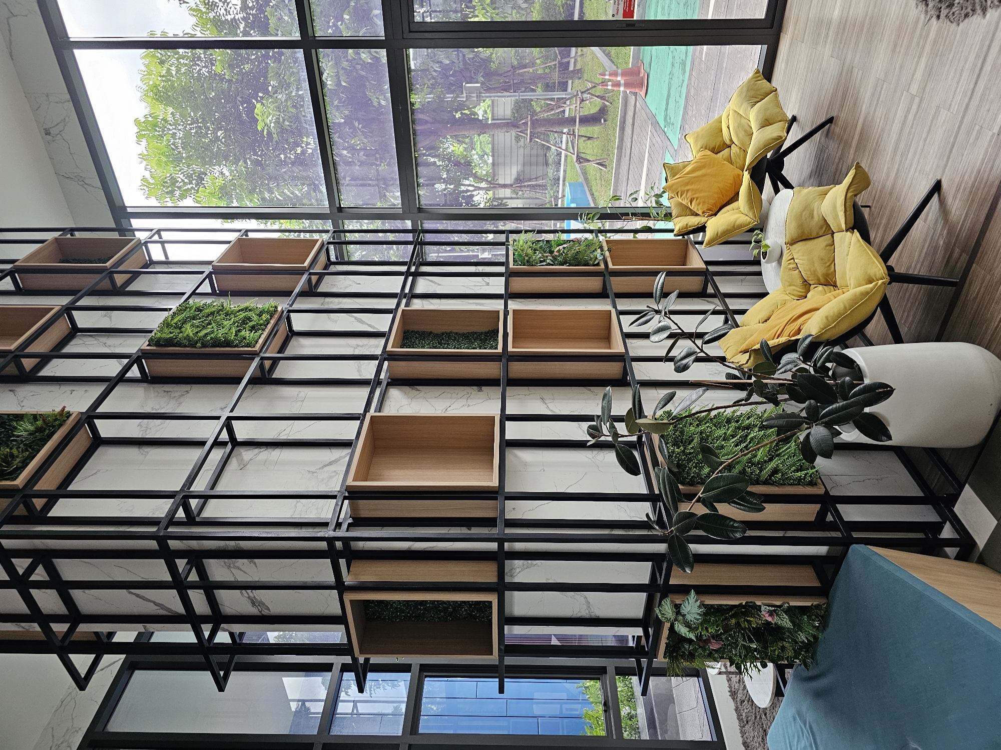 The image depicts a modern interior space with large windows overlooking an outdoor area. The interior features a tall, black metal shelving unit with numerous wooden cubbies, some of which contain artificial plants. The wall behind the shelving is decorated with marble-patterned tiles. In the foreground, there are two plush yellow armchairs with matching cushions placed near a small white circular table. A large potted plant with dark green leaves stands to the left of the armchairs. To the left of the shelving unit, a teal upholstered bench is partially visible. The outdoor view through the windows shows trees, a paved area, and orange traffic cones. The overall aesthetic suggests a contemporary lounge or lobby area within a building in Bangkok, Thailand, possibly a hotel or condominium. There are no people visible in the image.