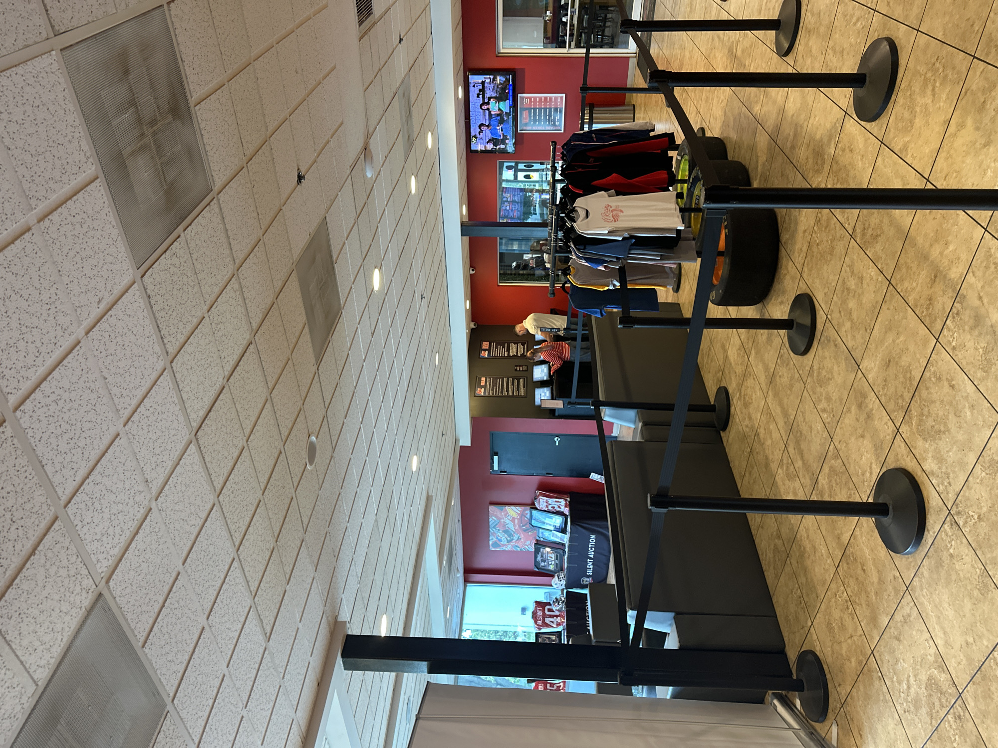 The image shows the interior of what appears to be a retail space in Orient Park, United States. There is a suspended tile ceiling with integrated lighting. The floor is tiled in a diamond pattern with a light beige color.
In the center of the frame, there's a merchandising area, displaying apparel on a rack and what appear to be racing tires for decoration. Adjacent to the display, black retractable barrier stanchions are set up, defining a queuing area. Further back, there is a counter with people, potentially staff and customers interacting. The background wall is painted red, featuring a television screen displaying content and framed signage. A closed door is seen in the background, alongside a table with merchandise related to a "Silent Auction." A large window provides a glimpse of the exterior.