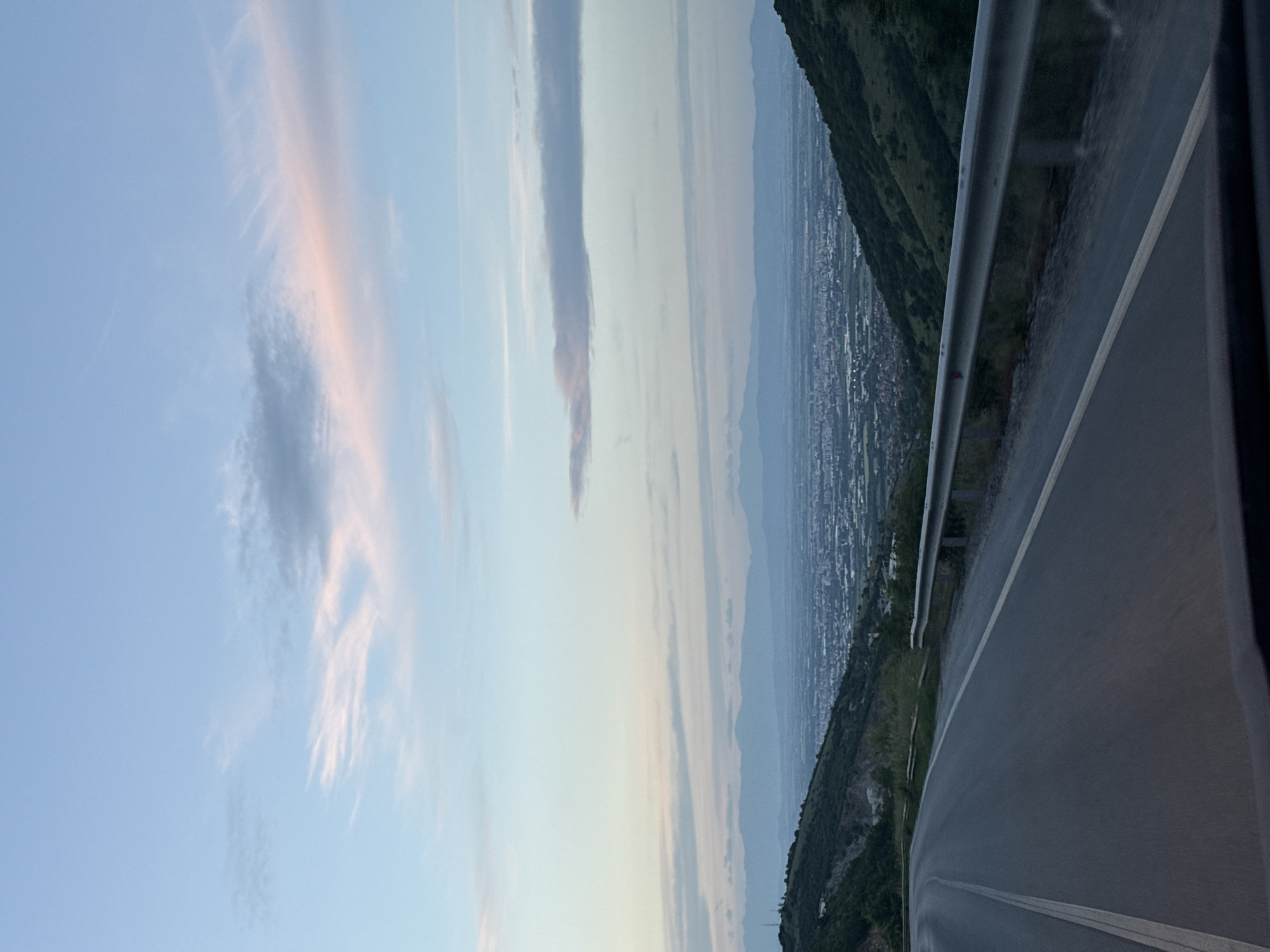 The image shows a view from a high road, likely in a mountainous area, overlooking a city. The location is likely in Belashtitsa, Bulgaria, given the context. The road has a grey metal guardrail. The road surface is dark grey asphalt. Beyond the road, there is a tree-covered hillside, and in the distance, a sprawling city extends to the horizon, possibly Plovdiv. The sky has pale blue and white clouds, some streaked with a pinkish hue. The perspective suggests the photo was taken from a vehicle.