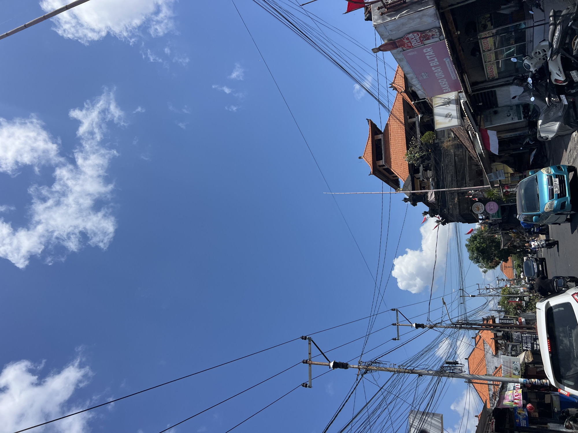 In Denpasar, Indonesia, on a sunny day, a street scene unfolds under a bright blue sky dotted with fluffy white clouds. Power lines crisscross the sky, suspended from utility poles that line the bustling street. Buildings with traditional Balinese architecture, featuring terracotta tiled roofs and intricate stone carvings, flank the road. Shops and eateries, some displaying vibrant signs, are visible along the roadside. A variety of vehicles, including cars, motorcycles, and scooters, navigate the street, indicating ongoing traffic. People are seen on motorcycles and walking on the sidewalks, suggesting daily life in the city. The overall atmosphere is one of a lively urban environment.