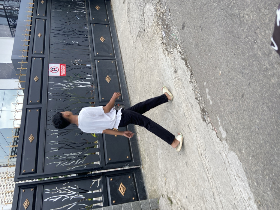 The image shows a young man walking on a paved road in front of a closed black gate with a "No Parking" sign. The gate is made of metal and has a decorative design of trees cut out in the middle. There are gold diamond-shaped accents on the gate. The man is wearing a white t-shirt, black pants, and white sandals. He is walking away from the camera and towards the right side of the image. The scene is located in Lembang, Indonesia, at coordinates -6.831 latitude and 107.607 longitude.