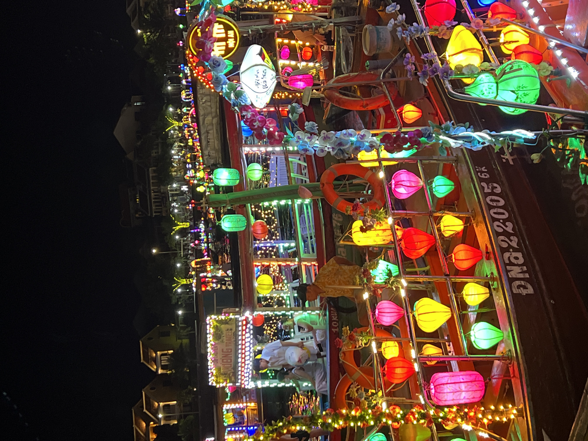 This image captures a vibrant night scene along the Thu Bon River in Hoi An, Vietnam, famous for its nightly lantern market. Several brightly lit boats are moored, adorned with numerous colorful lanterns of various shapes and sizes, including traditional Vietnamese silk lanterns. The boats are decorated with strings of white and colored lights, and floral garlands. People are visible on the boats, some seated and others standing, suggesting they are either passengers or vendors. The background shows the illuminated architecture of Hoi An's historic town. The atmosphere is festive and lively, highlighting the unique cultural charm of Hoi An at night.