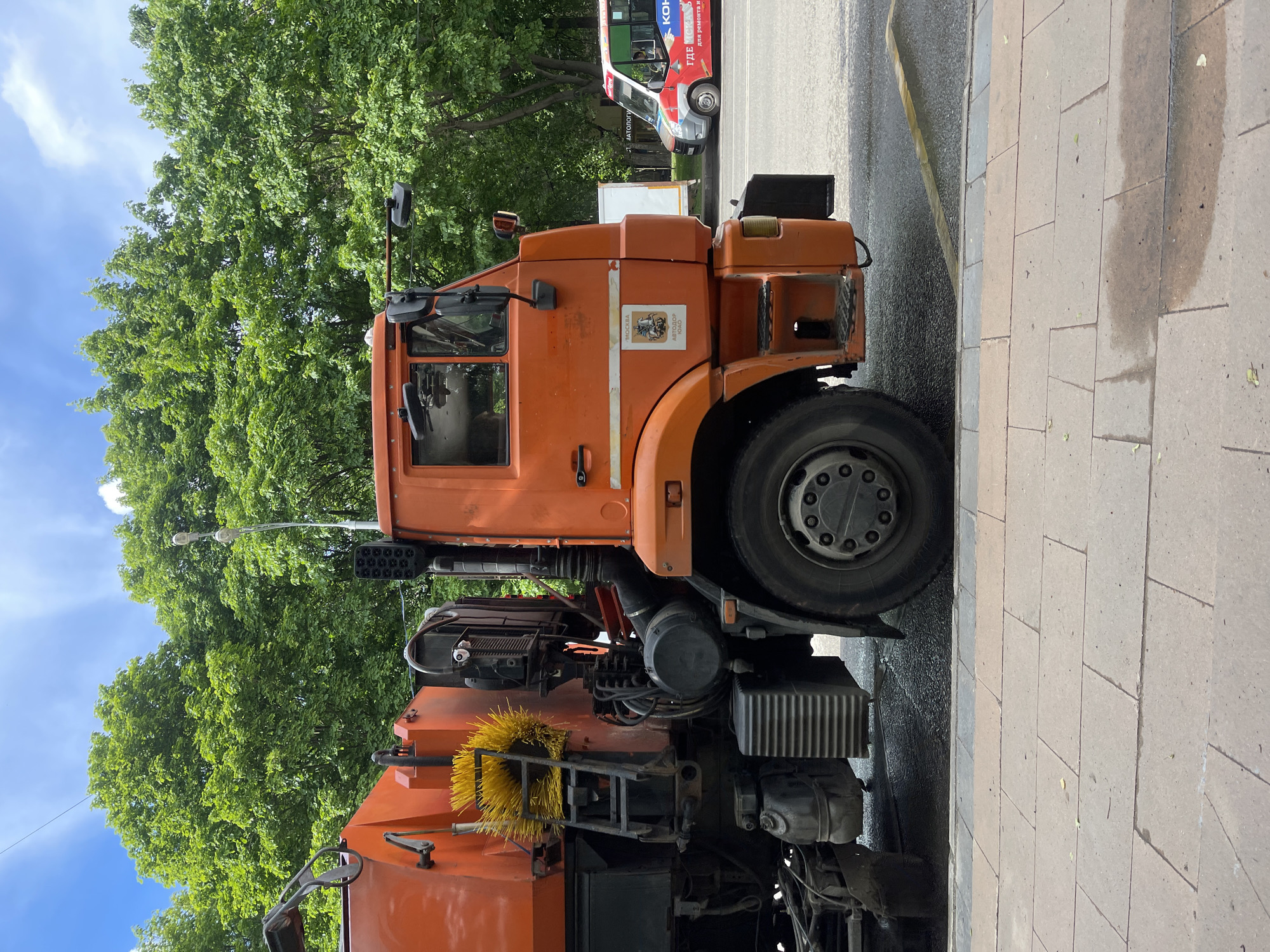 The image shows an orange street sweeper truck parked on the side of a paved road in Moscow, Russia. The truck has a distinctive emblem of the city of Moscow on its cabin. A section of pavement is visible next to the truck, and trees and a bus are in the background. The lighting suggests it is daytime.