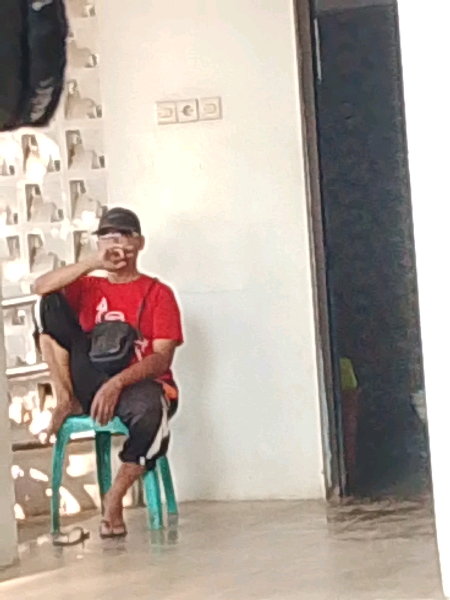A man wearing a red t-shirt, black shorts with white stripes, and a black baseball cap is sitting on a teal plastic stool. He is looking at the camera and has his right hand raised to his face. A black bag is slung across his chest. He is wearing sandals on his feet. The background appears to be a wall with a textured pattern and some electrical outlets. A dark doorway is visible on the right side of the image. The scene suggests an indoor setting, possibly a home or a public space within Probolinggo, Indonesia.