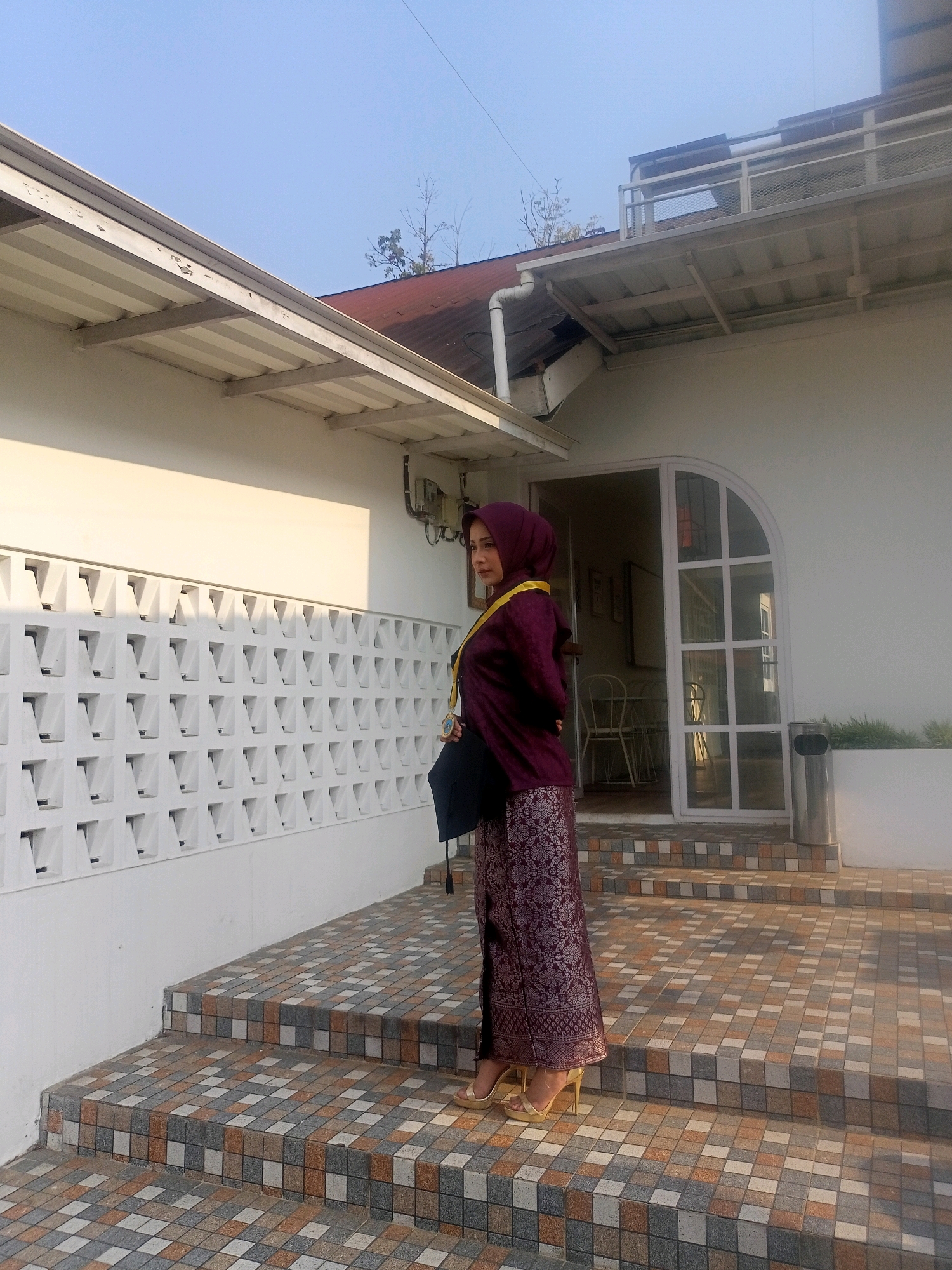 A young woman, wearing a maroon hijab and a patterned maroon skirt, stands on a tiled staircase outside a white building in Blambangan, Indonesia. She is dressed in formal attire, including a gold sash draped over her shoulders and high-heeled gold sandals. She holds a black bag in her left hand. The building features a white wall with decorative ventilation blocks, a white awning, and a large arched glass door. The staircase is covered in small, multi-colored tiles. The background shows a clear blue sky and a hint of a red-tiled roof. The lighting suggests it is daytime, possibly late afternoon due to the long shadows.
