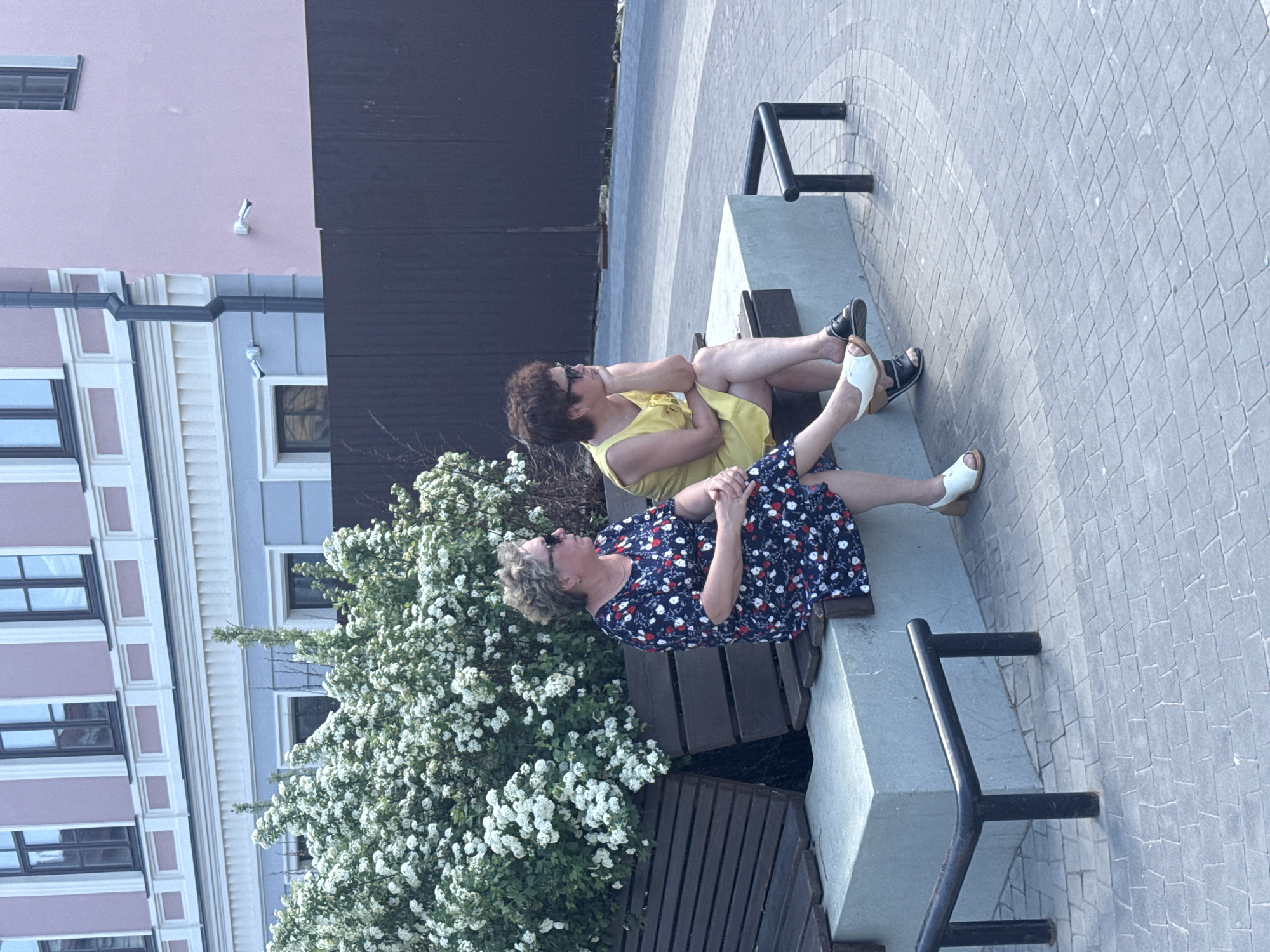 Two women are sitting on a park bench. One is wearing a yellow dress and sunglasses, while the other is wearing a blue and red floral dress and sunglasses. They are both sitting with their legs crossed. The bench is located in a public space with buildings in the background. There is a large bush with white flowers next to the bench. The location is Kazan, Russia. The ground is paved with stone tiles. There is a fence behind the bench.