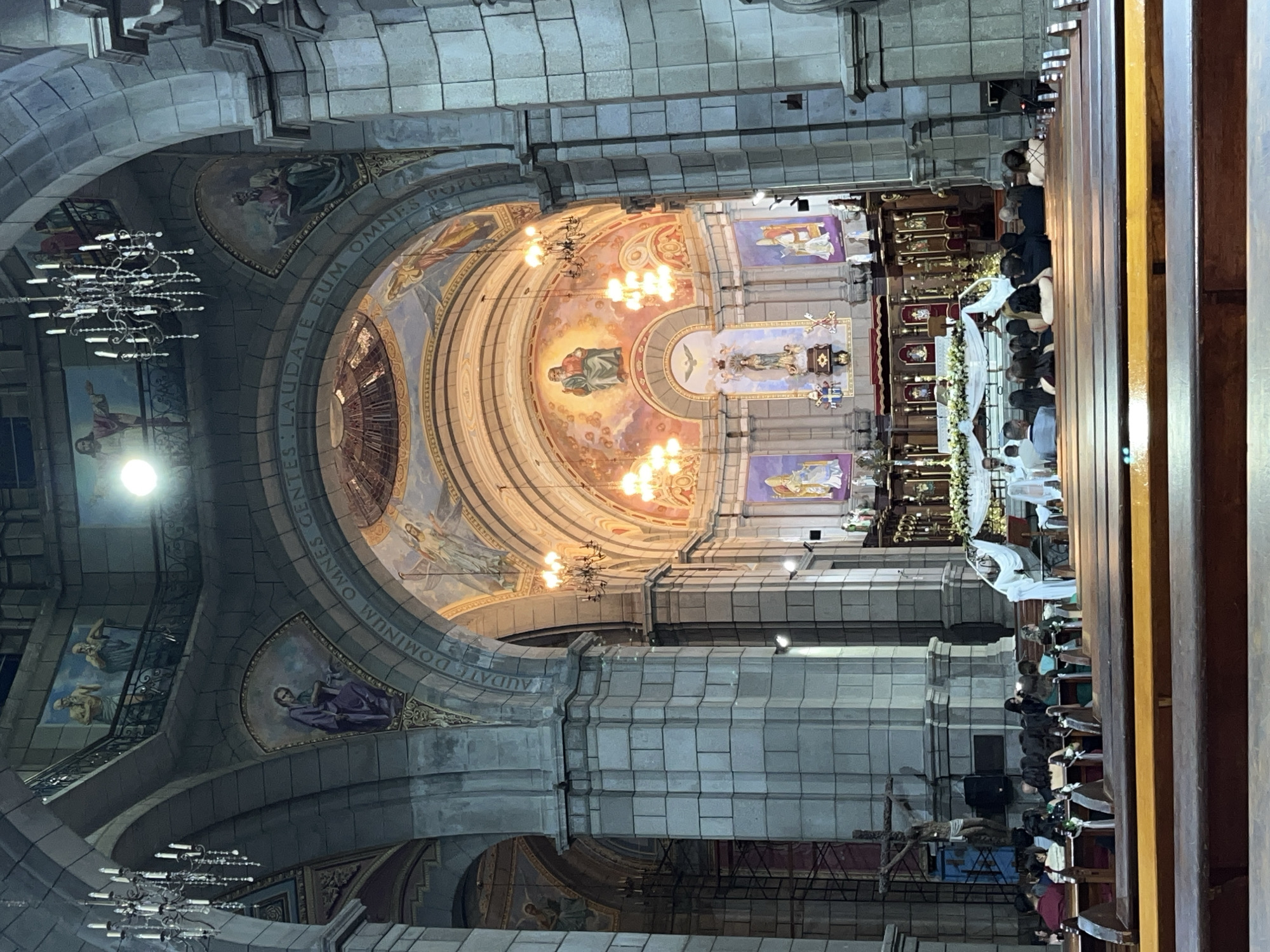 The image depicts the interior of the Mérida Cathedral in Mérida, Venezuela, during what appears to be a religious ceremony or service. The cathedral's architecture is grand, with large stone columns, arched ceilings, and ornate chandeliers. The walls and ceilings are adorned with religious paintings and frescoes, depicting biblical scenes and figures.
In the foreground, wooden pews are occupied by a congregation of people dressed in formal attire, facing the altar area. Some individuals are seated, while others are standing. The altar itself is decorated with flowers and draped in white fabric. A priest or clergyman, dressed in white vestments, is visible near the altar, possibly officiating the ceremony.
To the left of the altar, a crucifix is prominently displayed. The overall lighting suggests an indoor environment, with artificial lights from chandeliers and possibly spotlights illuminating the space. The scene conveys a sense of solemnity and reverence.