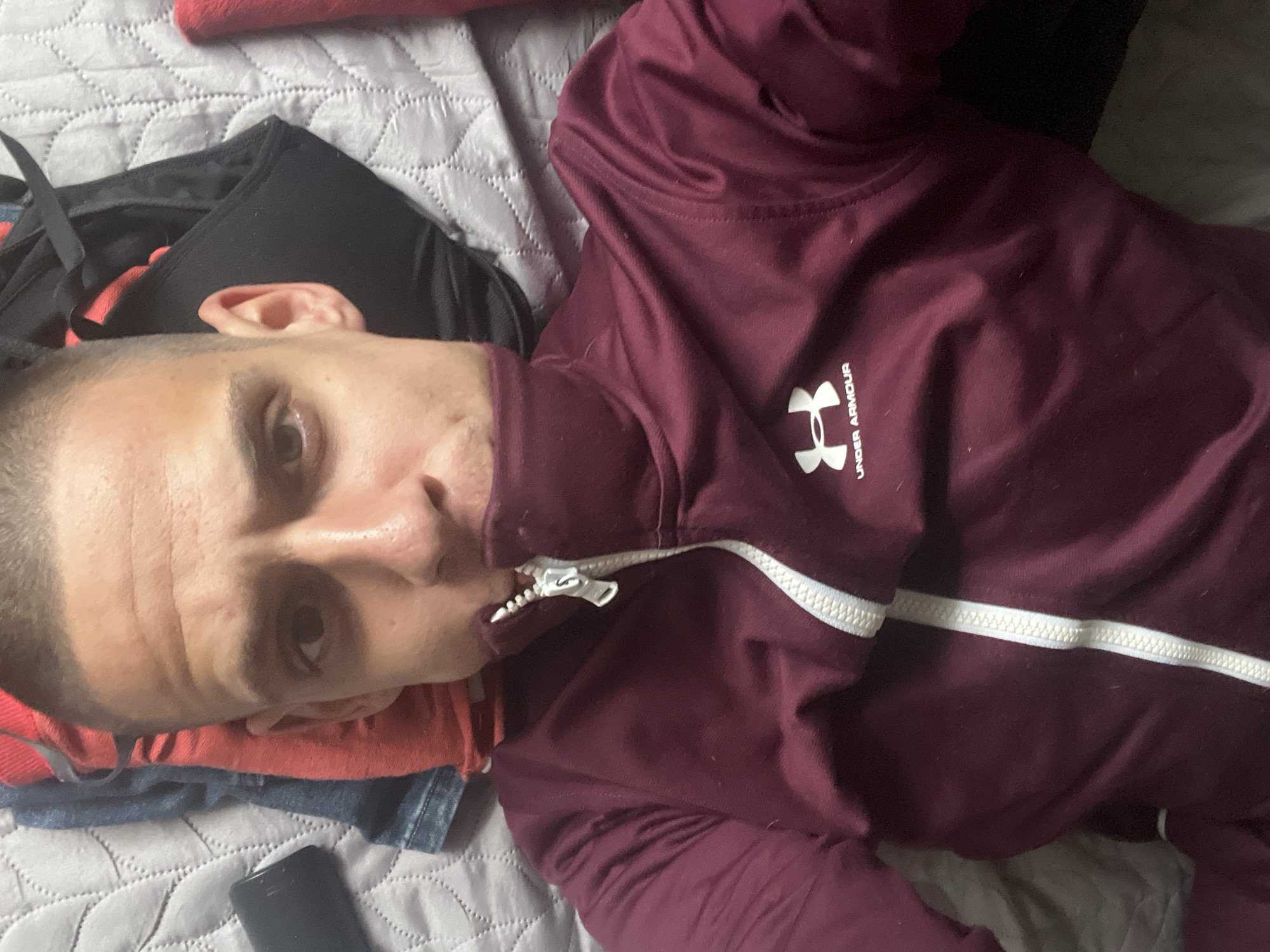 The image shows a man lying on a bed with a gray quilted bedspread. He is wearing a burgundy Under Armour track jacket with a white zipper. A black backpack and other clothing items, including what appears to be a red shirt and a piece of denim, are also on the bed. A black object, possibly a phone, is visible near the bottom of the frame. There is no indication of the location of Sofia, Bulgaria, in the image.