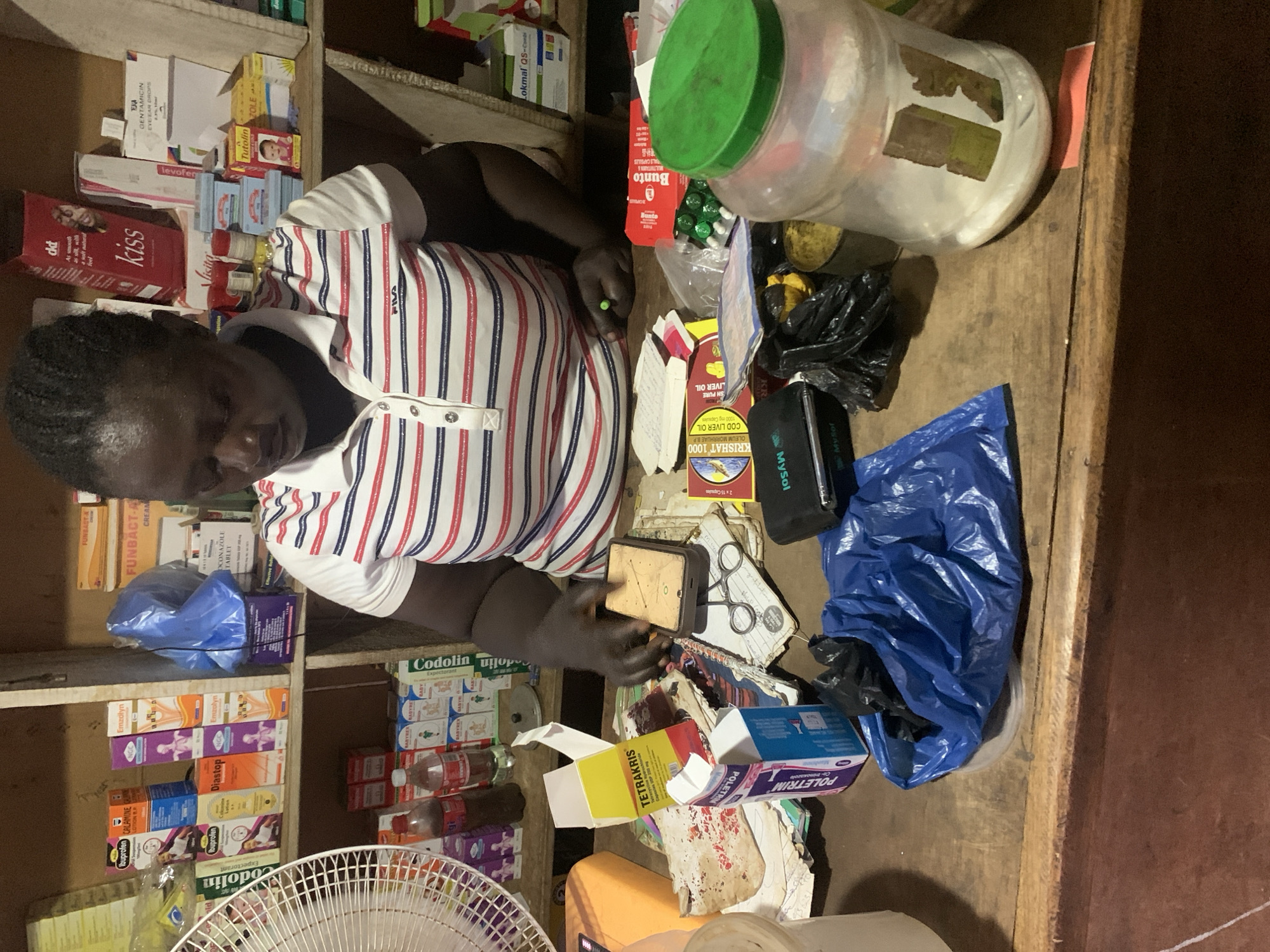 An overhead shot depicts a woman in Gbeleko, Nigeria, working at a counter in what appears to be a small pharmacy or dispensary. Shelves filled with various medications and health products are visible behind her. The counter is cluttered with items including containers of medications, boxes of drugs (TETRAKRIS, POLETRIM), bags, scissors, and miscellaneous papers. The woman, wearing a striped Fila shirt, is handling a sponge-like object. A partially visible electric fan sits in the lower left corner.
