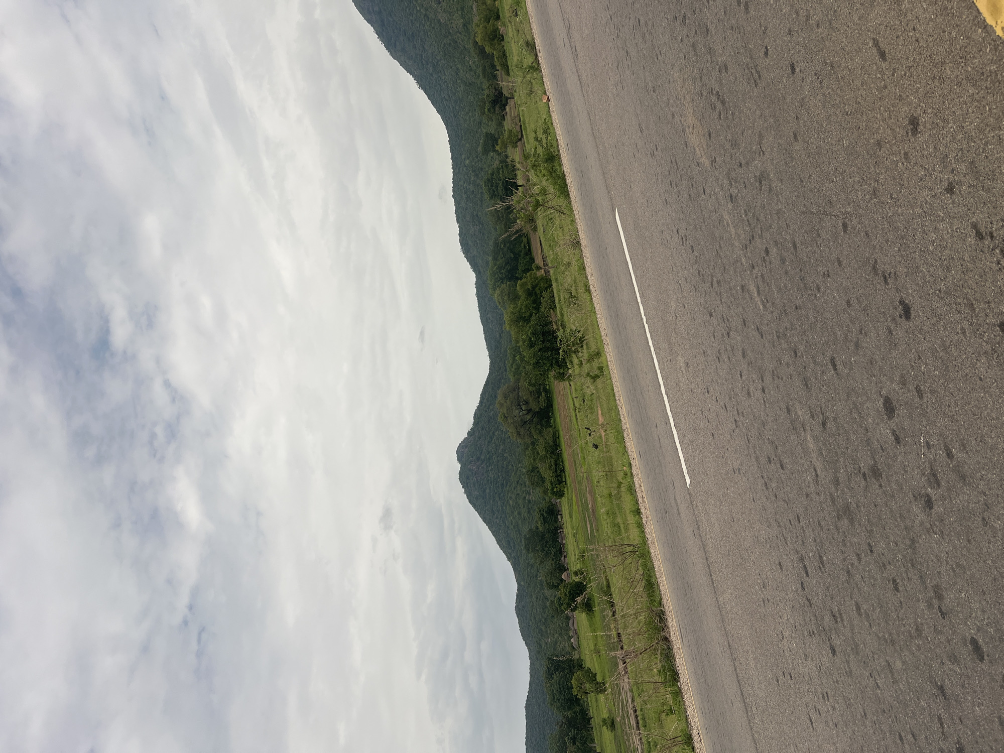 The image captures a road running alongside a lush, green landscape with rolling hills covered in dense vegetation. The road is paved and has a white line marking its edge. The sky above is overcast with dramatic, textured clouds. The scene suggests a natural, possibly rural setting in Minda, Nigeria. The perspective is from the side of the road, looking towards the hills and the sky. There are no people or specific objects immediately apparent, but the landscape itself is the primary subject. The time of day appears to be daytime, with the overcast sky creating diffused lighting. The overall mood is serene and natural, evoking a sense of travel or a journey through a verdant environment. There is no visible text in the image.