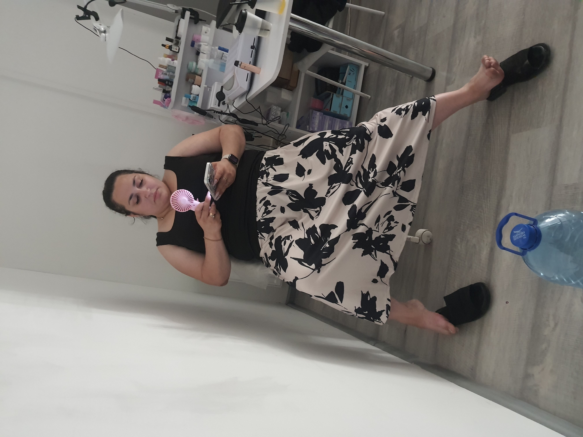 A woman is shown from the chest down, sitting with her legs extended, holding a small, pink, battery-operated fan. She is wearing a black tank top and a long, flowing skirt with a black and white floral pattern. Her left leg is bent at the knee, with her foot resting on a black sandal. Her right leg is straight, with her foot also in a black sandal. The woman appears to be looking down at the fan in her hands.
The setting appears to be indoors, possibly a salon or a treatment room, with a table of bottles and supplies visible in the background, along with a lamp. The floor is made of grey wooden planks. A large blue water bottle with a white cap is on the floor to the right of the woman. The lighting suggests it could be daytime. There is no discernible text in the image. The overall mood seems relaxed, possibly suggesting the woman is trying to cool herself down.
