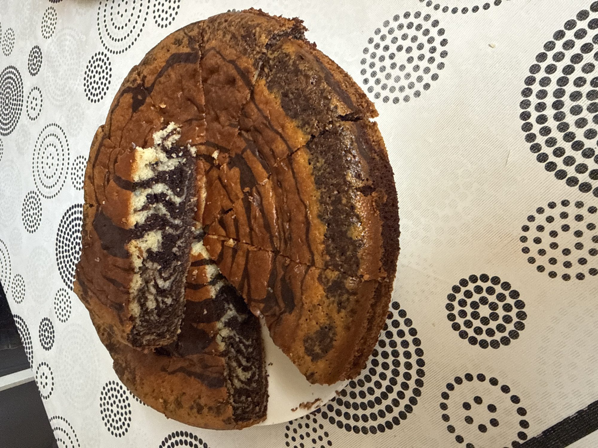 A whole marble cake is presented on a white plate, with one slice removed and placed on top of the cake. The cake has a swirled pattern of dark chocolate and lighter batter, and has been cut into multiple slices. The plate is on a white tablecloth with a repeating pattern of black circles made of dots. The scene is likely indoors, with a dark surface visible in the upper left corner, possibly a table. The lighting is natural and even.