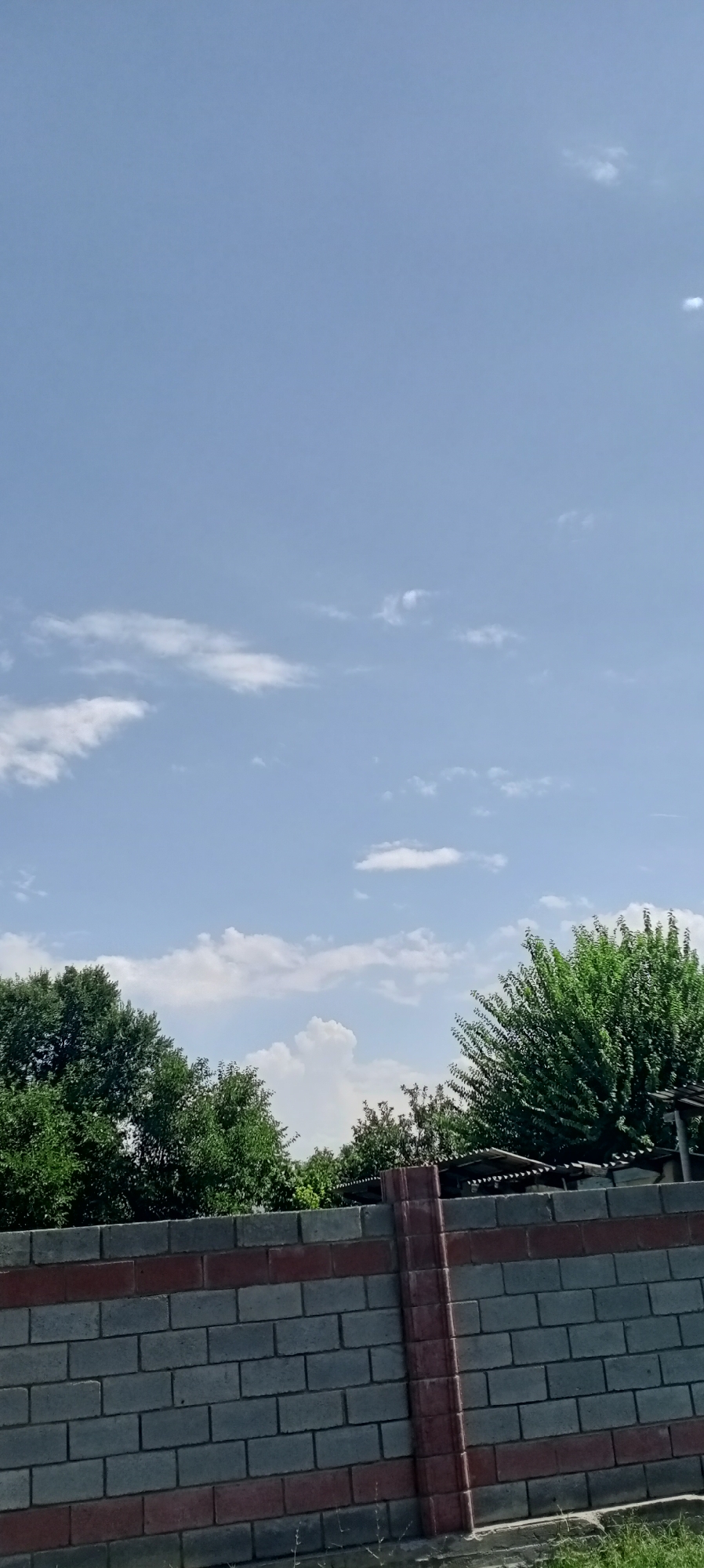 The image depicts a clear blue sky with scattered white clouds. In the foreground, a brick fence with red and grey bricks is visible. Behind the fence, lush green trees fill the lower portion of the frame. The scene appears to be an outdoor setting in the city of Sokuluk, Kyrgyzstan.
