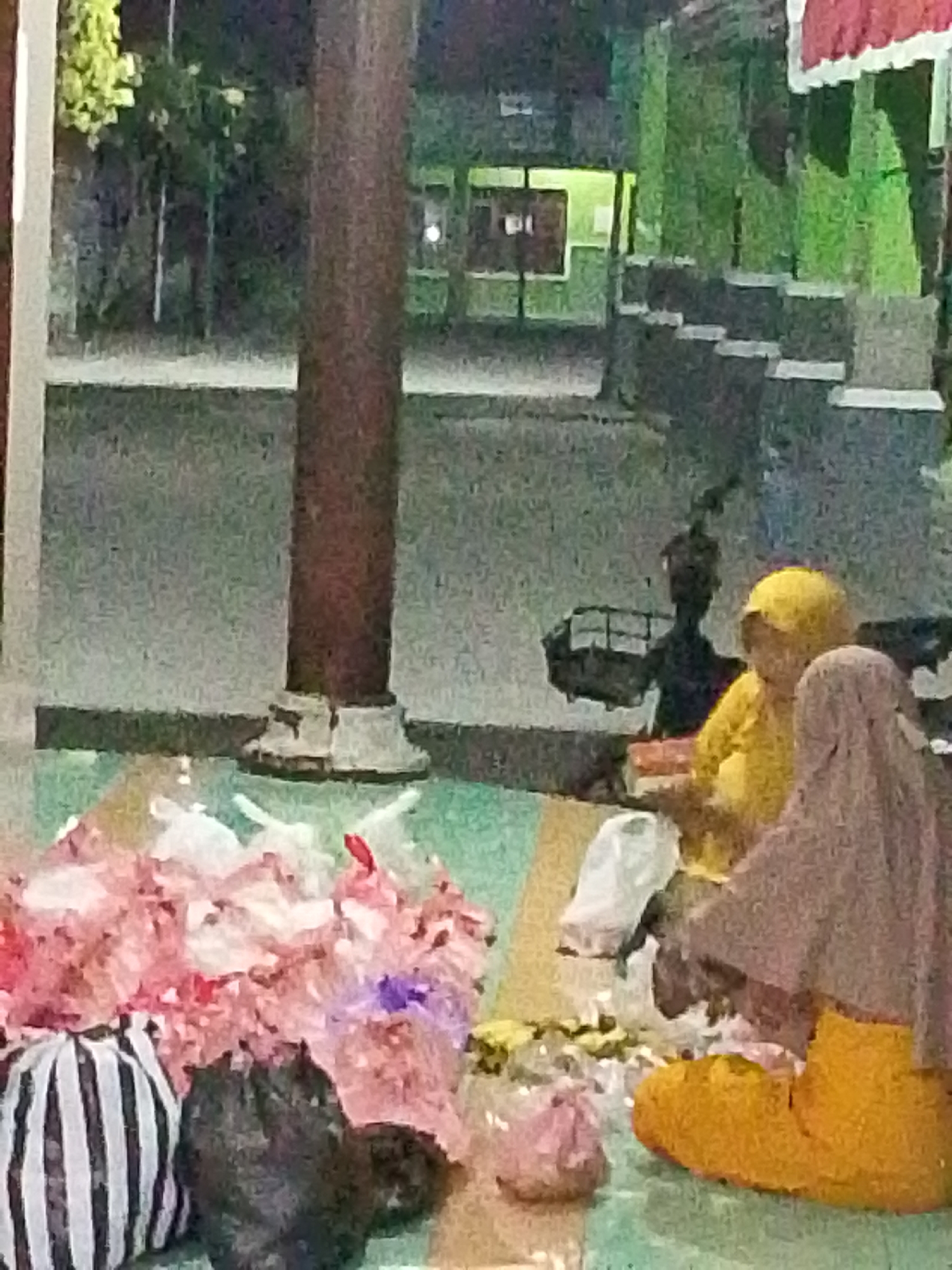 A group of people are gathered around a collection of colorful items on the ground, possibly in a courtyard or open space in Bulurejo, Indonesia. The scene appears to be at night, illuminated by artificial lights. In the foreground, there are several large bundles of what seem to be floral decorations, predominantly pink and white, with some purple accents. To the left of the floral arrangements, a large round object with black and white stripes is visible. Next to it is a dark, amorphous object, possibly a bag or another decorative item.
Two individuals are seated in the mid-ground. One person wears a bright yellow head covering and a yellow garment. Another person is draped in a light brown or grey hijab and a bright orange garment on their lower body. They appear to be interacting with the items on the ground. In the background, a large, dark, vertical pillar dominates the center of the frame. Behind the pillar, a building with green walls and what appears to be illuminated windows can be seen. The ground surface is a textured mix of green, yellow, and white, suggesting it might be a tiled or painted floor. The overall atmosphere is subdued due to the low light conditions, with some areas in shadow and others brightly lit.
