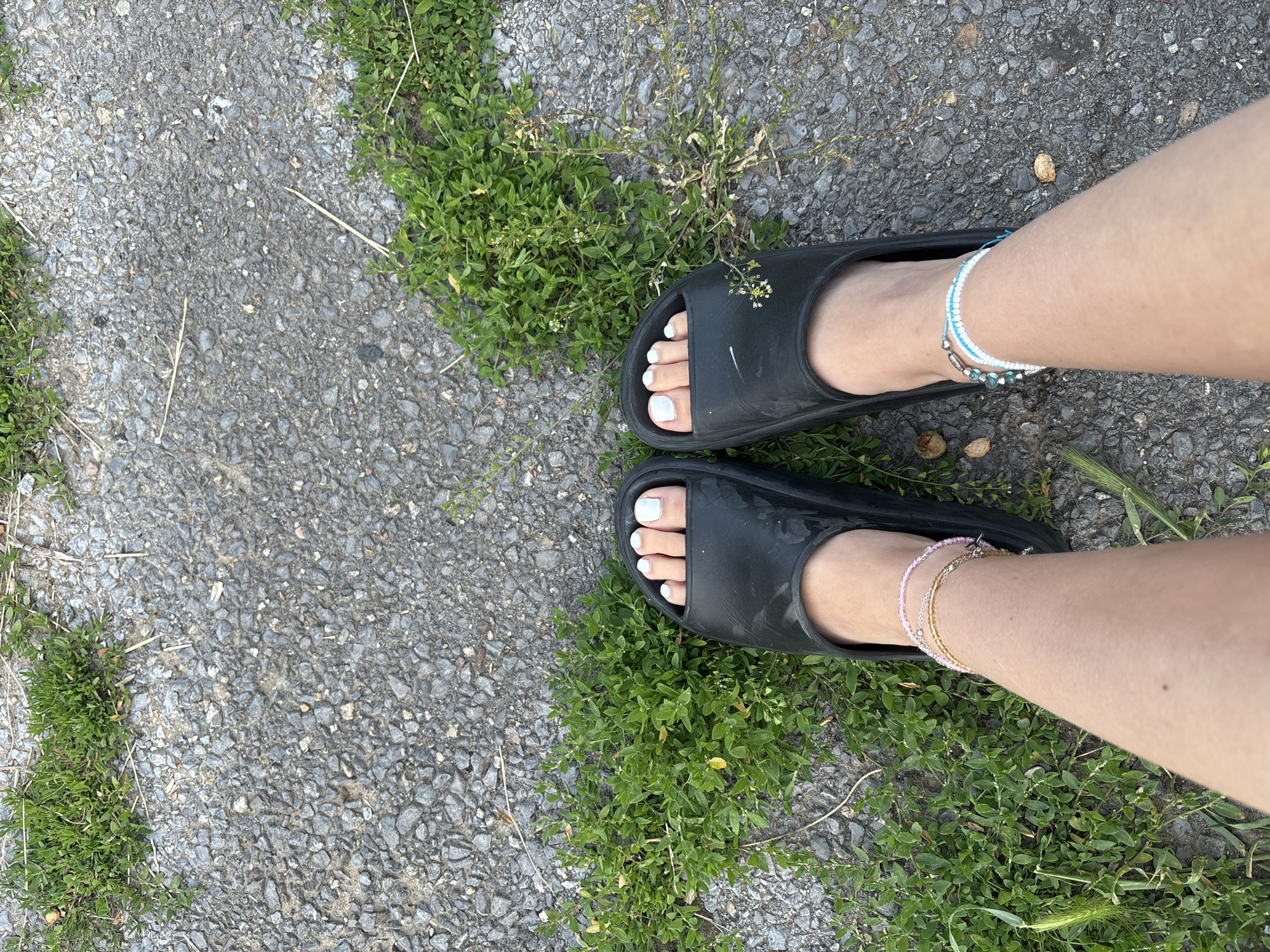 The image shows a first-person perspective of a person's feet wearing black slide sandals. The toenails are painted a light blue-white color. There are two beaded ankle bracelets on the person's left leg, one with pink and gold beads, and another with white and blue beads on the right leg. The person is standing on a textured gray surface, possibly asphalt or concrete, with patches of green grass and small weeds growing around the feet. The lighting suggests it is daytime. The scene appears to be outdoors, and the natural elements like grass and textured ground suggest a park, sidewalk, or unpaved path. The image does not provide any specific visual cues to place it within the city of Sofia, Bulgaria.