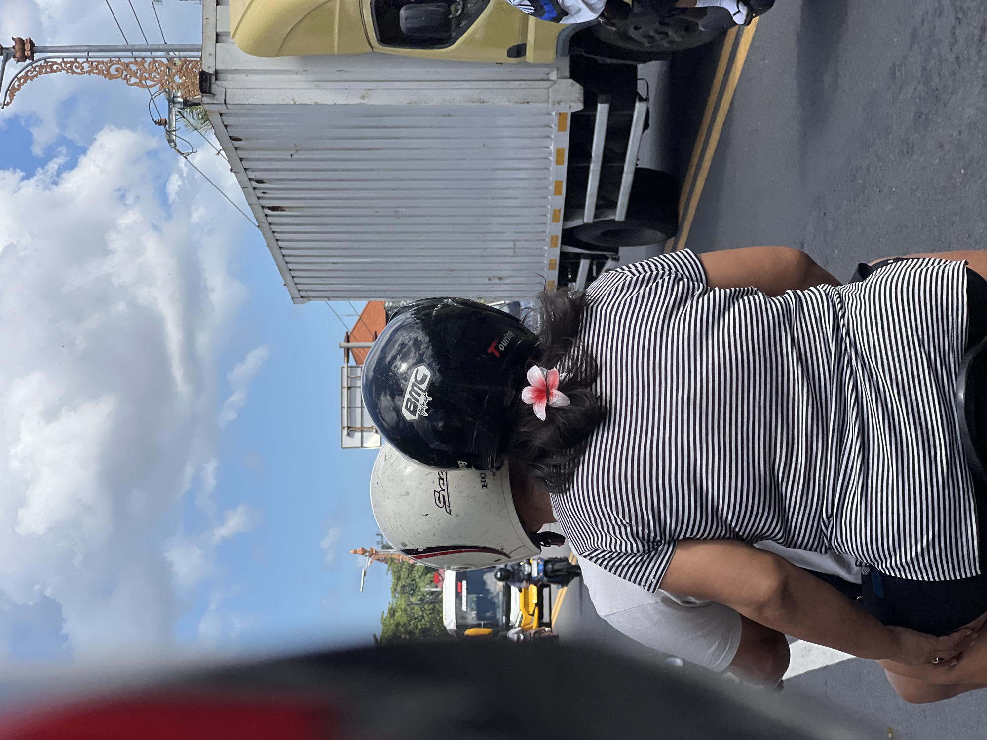 A rear view of two people on a motorcycle in Denpasar, Indonesia. The person in the front is wearing a white helmet with a black logo and the person in the back is wearing a black helmet with a pink flower in their hair. They are both wearing casual clothing and are on a road with a yellow line on the right side of the road. There is a large white truck on the right side of the road and a yellow bus in the distance on the left side of the road. The sky is blue with some white clouds.