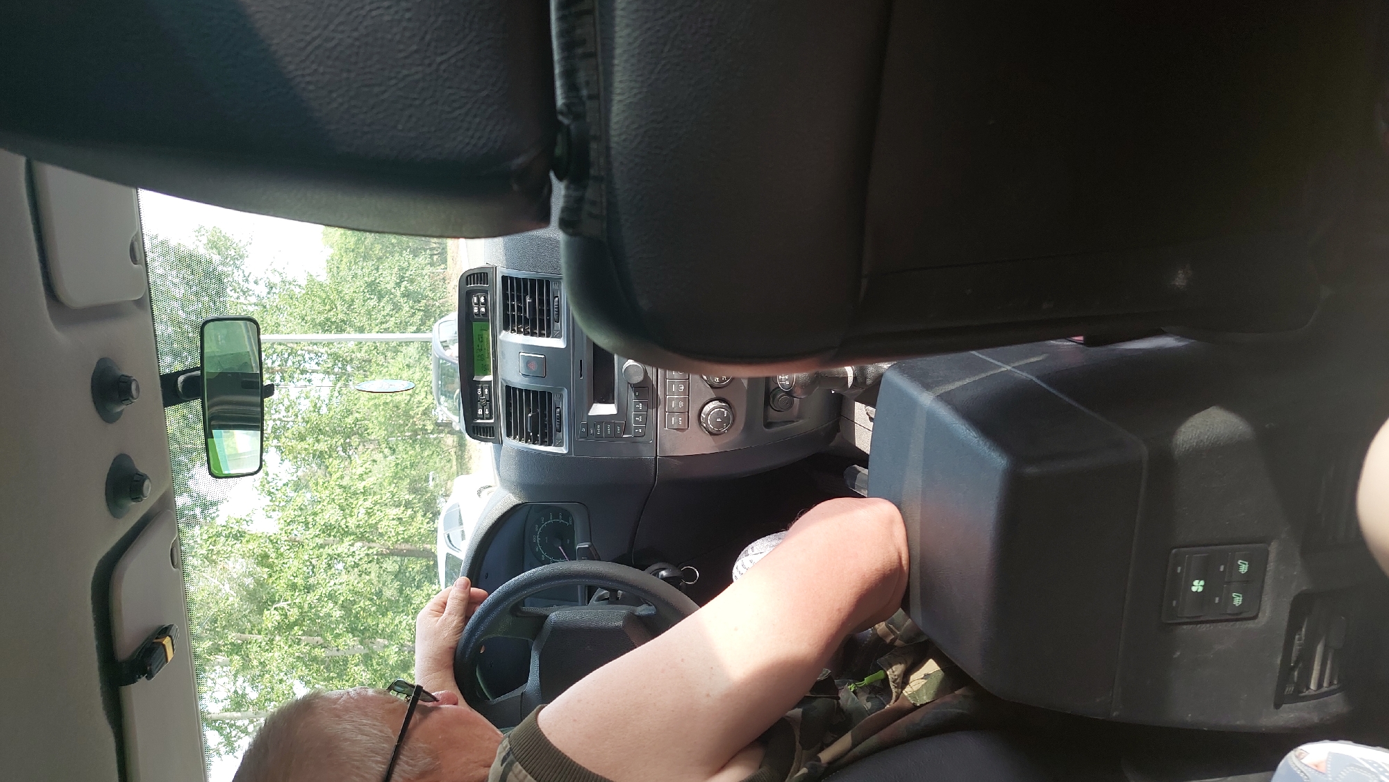 The image shows the interior of a car, likely a SUV, from the perspective of a passenger in the back seat. In the foreground, the back of a dark, leather-upholstered headrest and seat is visible. To the left, a man with graying hair and glasses is seated in the driver's seat, his left hand resting on the steering wheel. He is wearing a camouflage-patterned shirt. The car's dashboard is visible, featuring a radio, air vents, climate control buttons, and a gear shift. A center console with buttons and a storage compartment is also prominent. The interior appears to be relatively modern. Outside the car, through the windshield and side windows, lush green trees and foliage are visible, suggesting a natural or suburban setting. The overall lighting indicates it is daytime, with sunlight filtering into the car. The image is cropped in a way that the view of the road and other vehicles is limited. The location is specified as Amursk, Russia, but there are no specific visual cues in the image to confirm this.