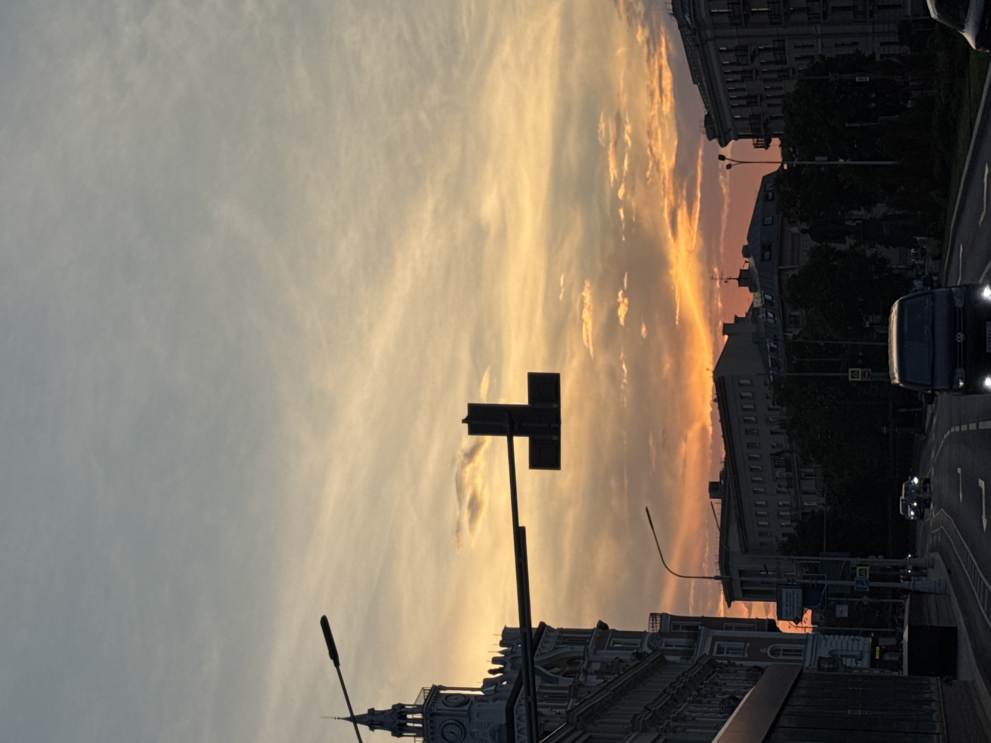 The image depicts a street scene in Moscow, Russia, during sunset. The sky is filled with vibrant hues of orange and yellow, indicating the sun is low on the horizon. In the foreground, a dark-colored van is driving on a paved road. Several other vehicles are also visible on the road.
Several buildings line the street, with varying architectural styles. One building on the left features a clock tower and ornate details. Another building on the right appears to be a multi-story residential or office building. Streetlights and traffic signals are present, illuminating the road. Trees are visible, lining parts of the street and the sidewalks. The overall impression is of a city transitioning from day to night, with the warm glow of the sunset casting a dramatic light on the urban landscape.
