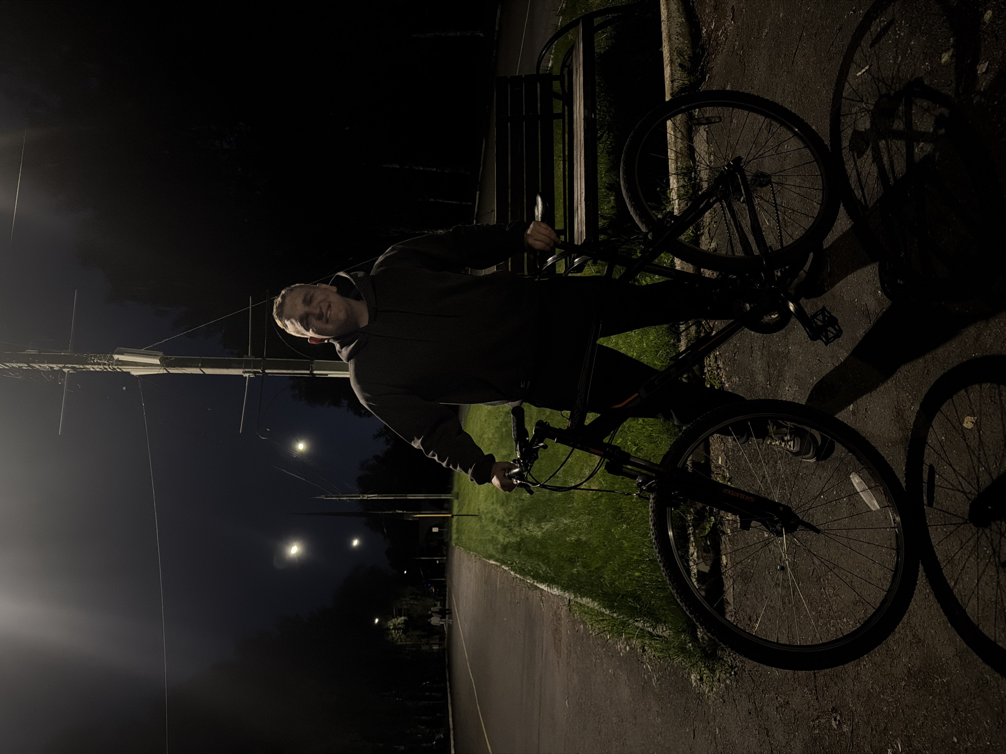 The image, rotated 90 degrees clockwise, captures a young man standing with a bicycle on a paved path at night. The man, appearing to be in his late teens or early twenties, is positioned in the center, wearing a dark hoodie and dark trousers. He is smiling warmly at the camera, holding the bicycle's handlebars with his left hand.
The bicycle, a dark-colored mountain bike with front suspension, occupies the right side of the frame. The text "SR SUNTOUR" is visible on its front fork. The bicycle casts a long, distinct shadow onto the dark asphalt path, illuminated by artificial light sources.
The setting is an outdoor area, likely a park or a recreational path in Izhevsk, Russia, during nighttime. The dark sky is punctuated by several bright streetlights, one of which creates a strong backlight and reflections on utility wires in the upper left. Utility poles are also visible further back. A strip of well-lit green grass borders the paved path. A dark wooden bench is partially visible behind the man and his bike. The overall mood is casual and pleasant, indicated by the man's smile. The clear sky suggests fair weather.