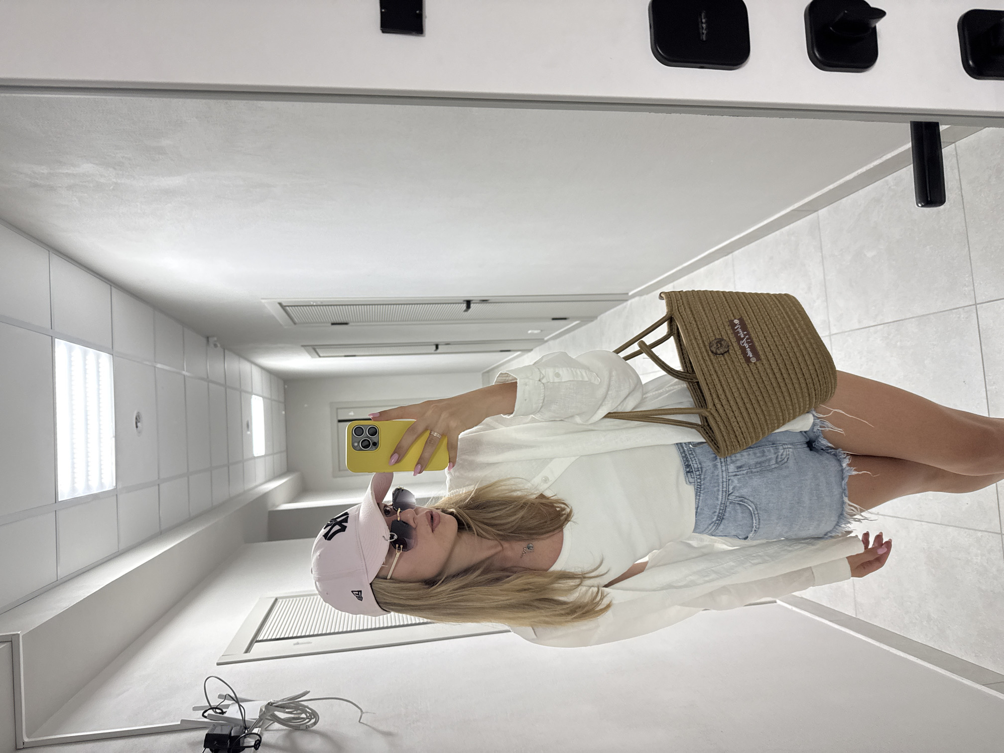 A woman with blonde hair, wearing a pink baseball cap, sunglasses, a white tank top, an unbuttoned white shirt, and denim shorts, takes a selfie in a hallway. She is holding a yellow smartphone in her right hand and a woven brown bag in her left. The hallway has white walls, tiled floors, and several doors. The lighting appears to be from overhead fluorescent panels. The scene suggests an indoor, possibly residential or hotel, environment.