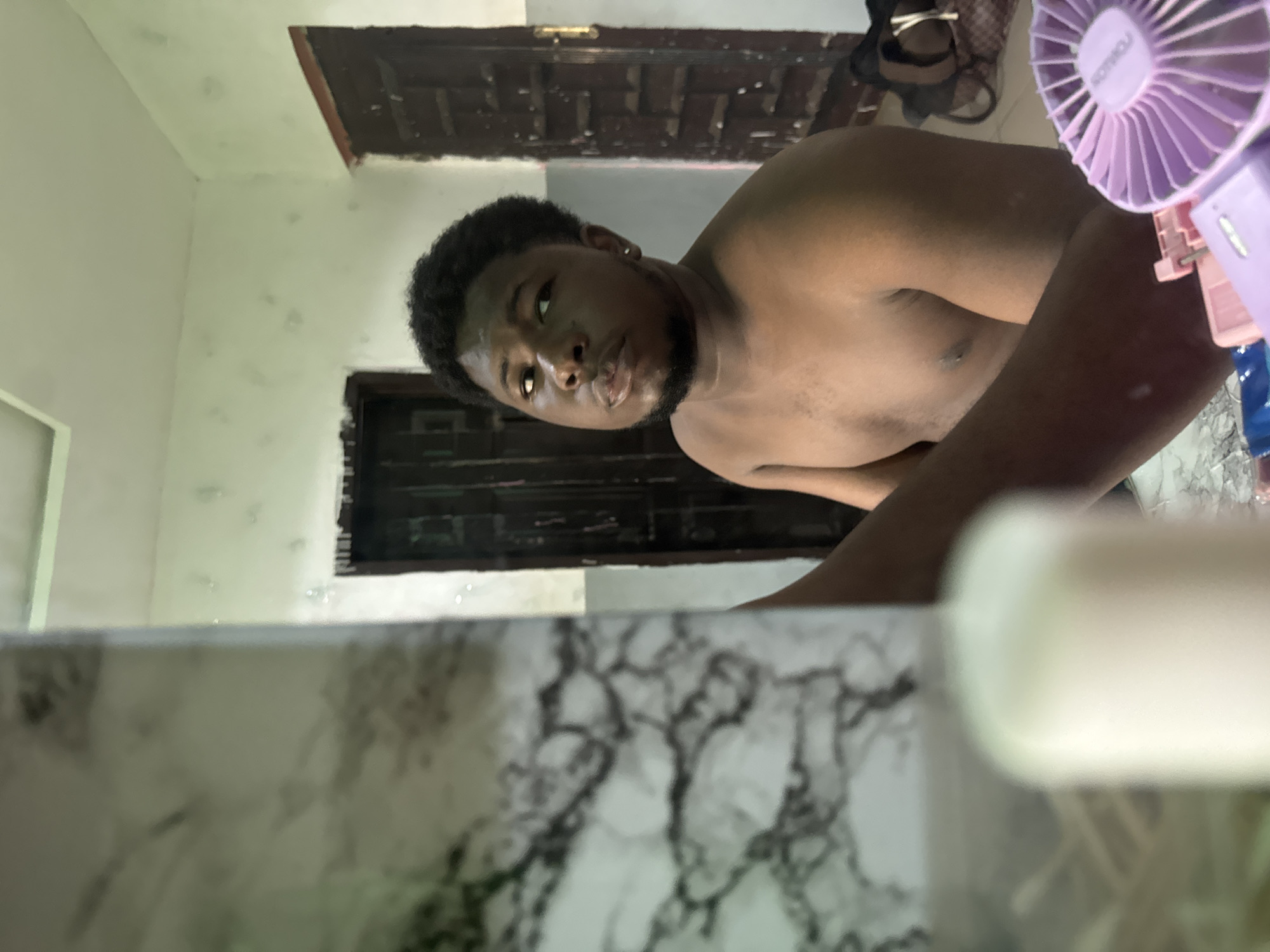 A shirtless man with dark hair and a beard is looking at the camera. In the foreground is a white and gray marble surface with some blurred white objects. In the background are two dark doors. In the top right of the photo is a purple fan. There are some shoes visible on top of one of the doors. The photograph seems to be taken indoors and may possibly be a bathroom. The location is unknown.