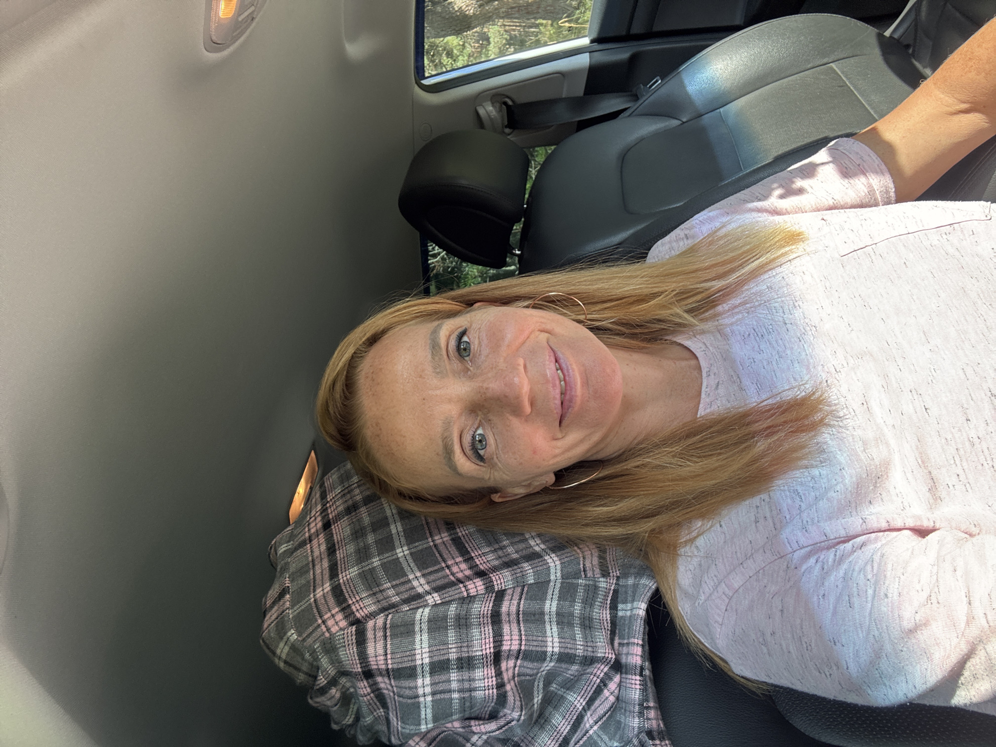 In Portovoe, Russia, a woman with blonde hair and freckles, wearing a pink t-shirt, takes a selfie from the passenger seat of a car. Her hair is shoulder-length and slightly windswept, and she is wearing hoop earrings. In the background, a black car seat and a gray and pink plaid blanket are visible. Outside the car, trees can be seen through the window, suggesting a natural, possibly rural, setting. The lighting indicates it is daytime, with sunlight casting shadows and highlights on the woman's face and clothing.