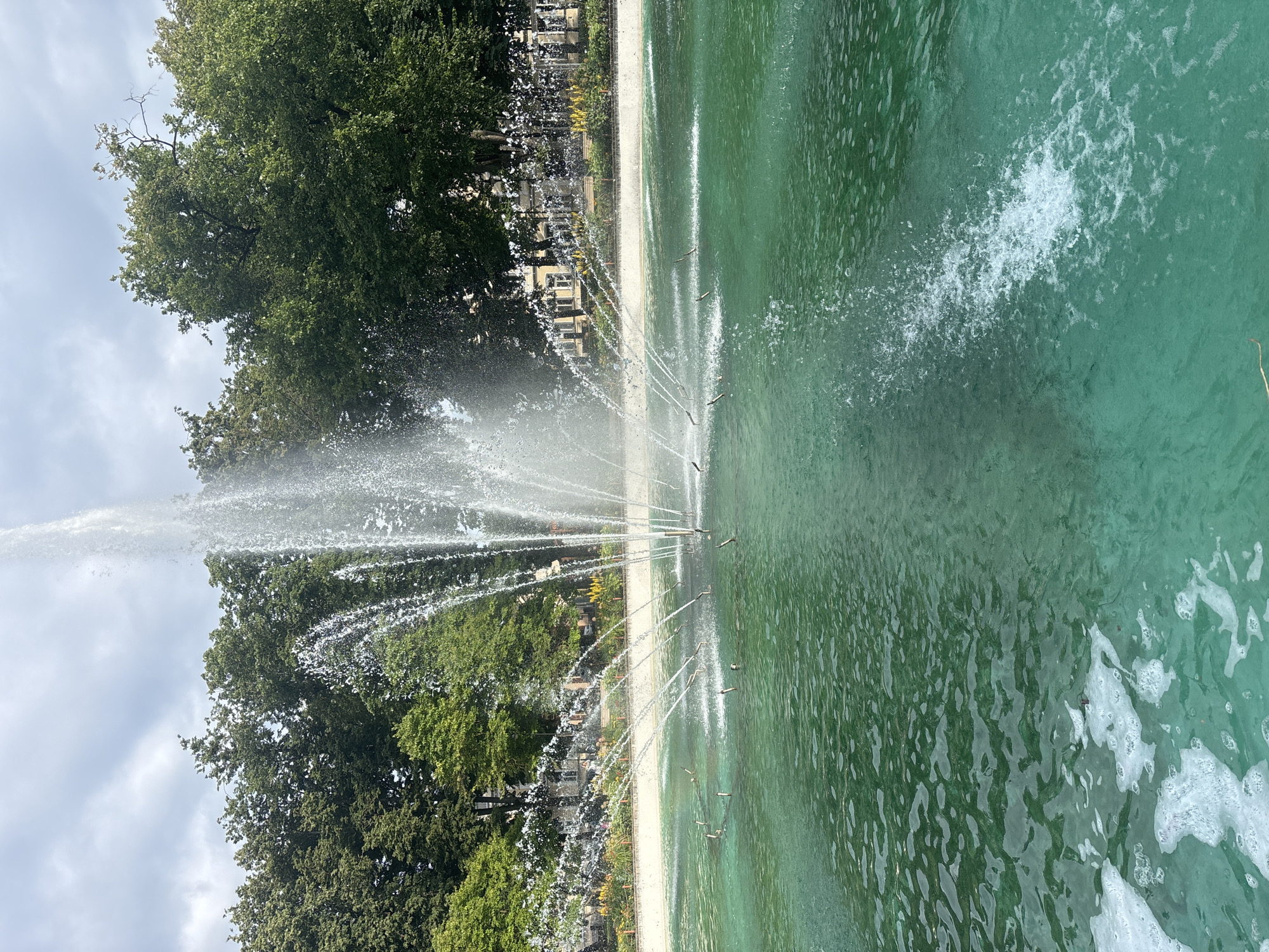 A wide shot captures a large, multi-tiered fountain in a park-like setting. The fountain's water is a vibrant green, with white foam gathering on the left side of the frame. Water jets are actively spraying upwards and outwards, creating a dynamic effect. In the background, a line of lush green trees forms a natural canopy. Behind the trees, a few buildings with classical architecture are visible, suggesting an urban environment. The sky above is overcast with scattered clouds. The scene is one of a public park with a prominent water feature, likely a place for leisure and relaxation. The image does not provide specific clues to identify its location within Daugavpils, Latvia.