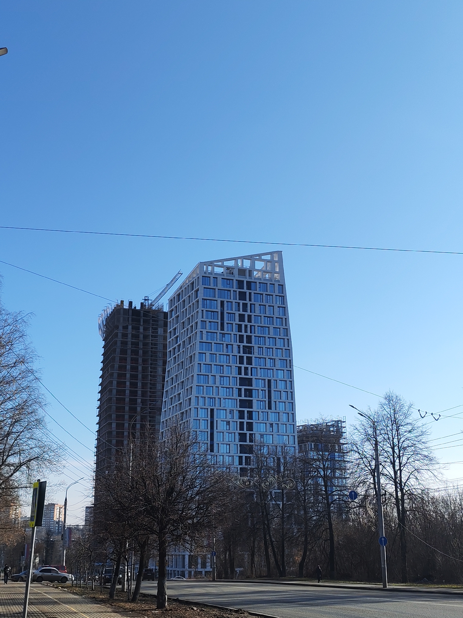 The image depicts a street scene in Izhevsk, Russia, with two tall buildings dominating the background. The building on the right is a modern high-rise with a white facade and numerous windows. The building on the left is under construction, its unfinished concrete structure visible. A crane sits atop this building. Wires crisscross the sky above the buildings and trees.
In the foreground, a street with a sidewalk and sparse trees runs along the side of the street. There are cars parked along the sidewalk, and pedestrians are walking on the sidewalk. The sky is clear and bright blue. Bare trees line both sides of the street. The light suggests it is daytime, possibly late morning or early afternoon. A street lamp is also present.