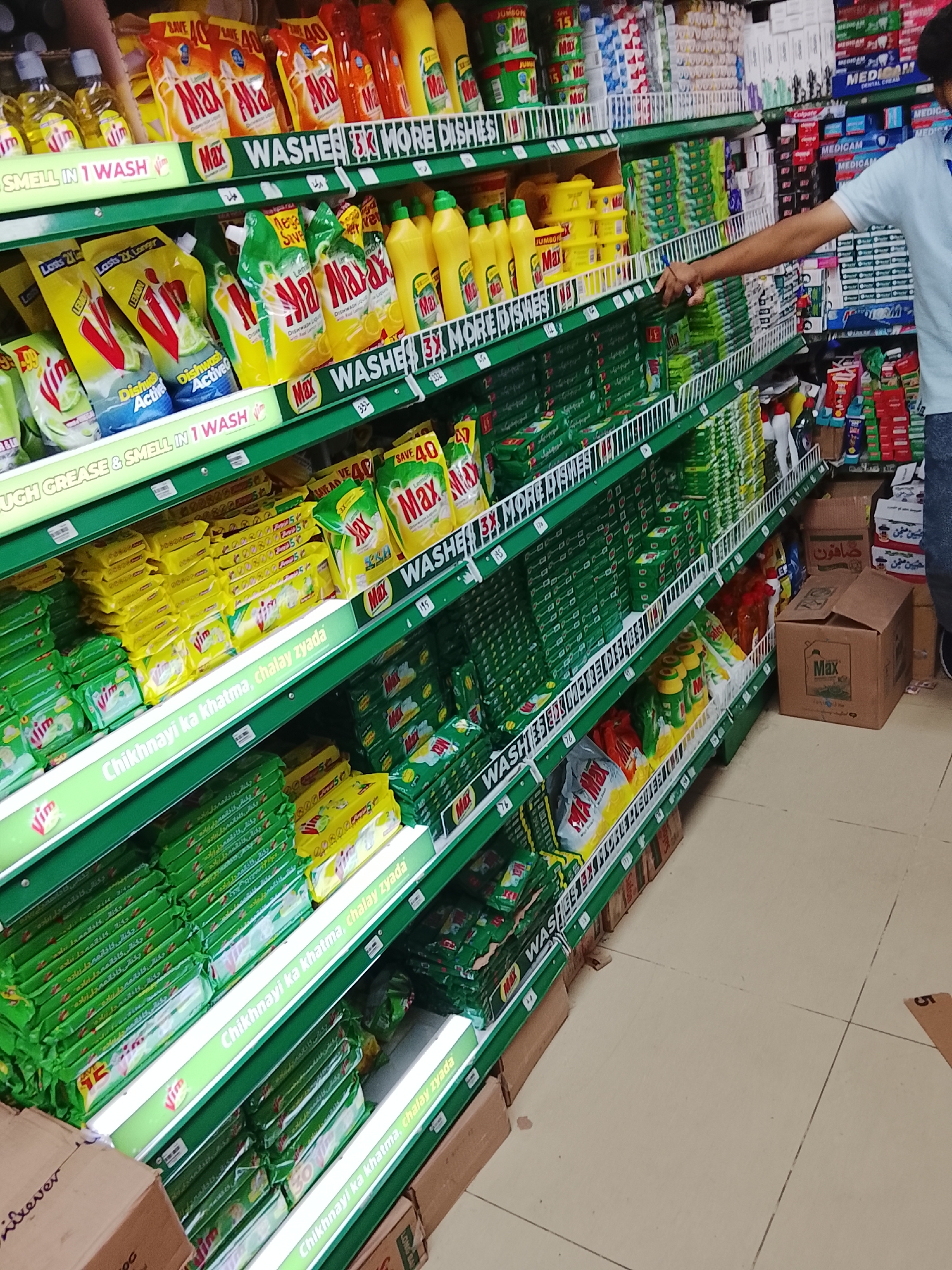 The image shows a person standing in an aisle of a store, possibly in Gulgasht Colony, Pakistan, as many of the products have Urdu text. The shelves are stocked with dishwashing detergents and soaps, primarily brands like "Max" and "Vim". A wide variety of cleaning supplies are placed on the green shelves. The person is reaching for one of the products. Other items such as toothpaste are on the shelves in the background. The floor is tiled. There are boxes of products near the base of the shelves.