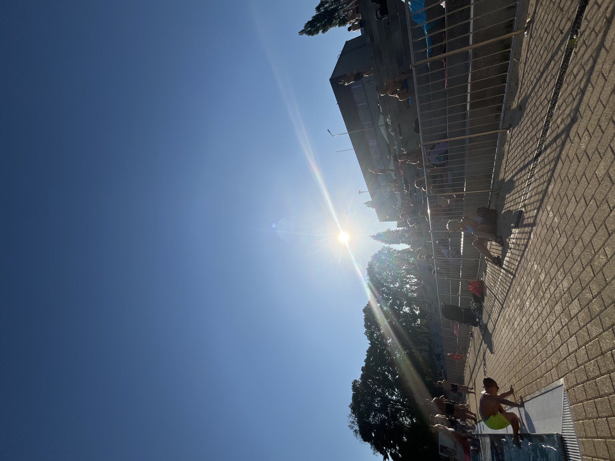 The image depicts a sunny day at an outdoor swimming pool, likely a diving platform area. The sun is bright and high in the sky, creating lens flare and casting long shadows on the brick-patterned ground. A metal railing lines the side of the pool, separating it from the surrounding area where several people are visible.
The main subjects are people, many of whom appear to be young men and boys. They are dressed in swimwear and are engaged in various activities. Some are sitting or standing near the pool's edge, potentially waiting for their turn to dive or swim. Others are shown in mid-action, possibly jumping or preparing to jump from the diving platform. One individual in the foreground is seated on the edge of the pool, looking towards the water.
The scene is bustling, with a crowd of people gathered, suggesting a recreational event or a popular swimming spot. The clear blue sky and bright sunlight indicate a warm, fair-weather day. The architecture of a multi-story building is visible in the background, alongside trees and vegetation, suggesting an urban or suburban setting. The overall mood seems lively and energetic, typical of a public swimming pool on a sunny day. No visible text is present in the image.