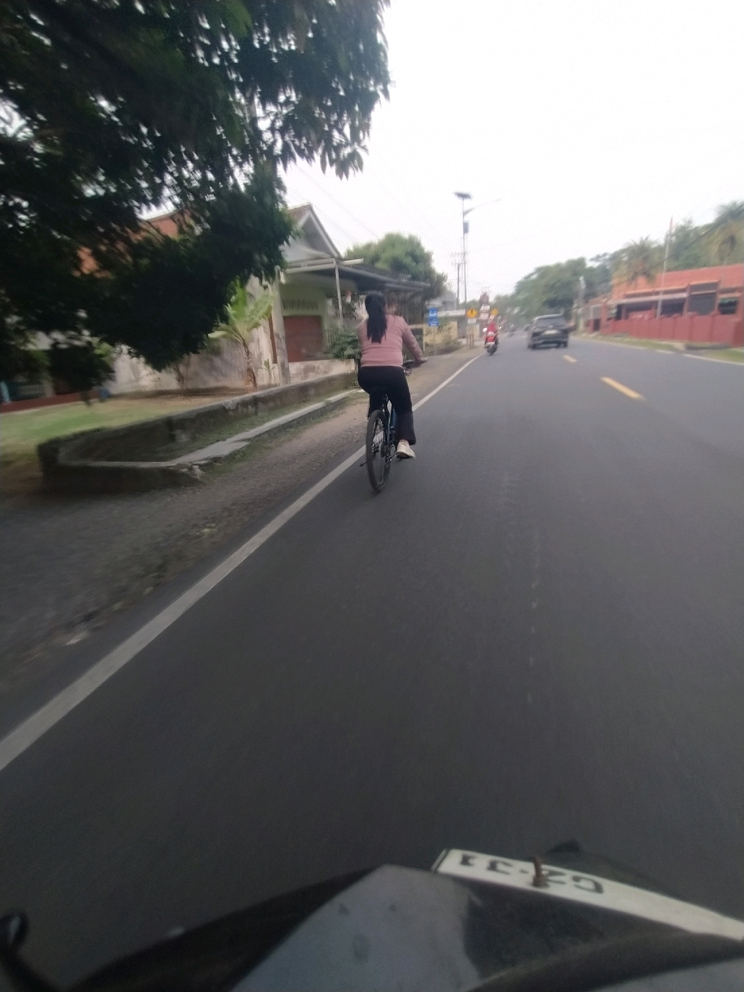 A person is riding a bicycle on the side of a road in Desa Karangbenda, Indonesia. The cyclist is wearing a pink long-sleeved shirt and black pants. The road is paved with asphalt and has a white line marking the lane. On the left side of the road, there is a ditch with some vegetation. Houses are visible in the background. A red motorcycle is riding in front of the cyclist, and a black car is further down the road. The sky is overcast. The image appears to be taken from the perspective of another vehicle.