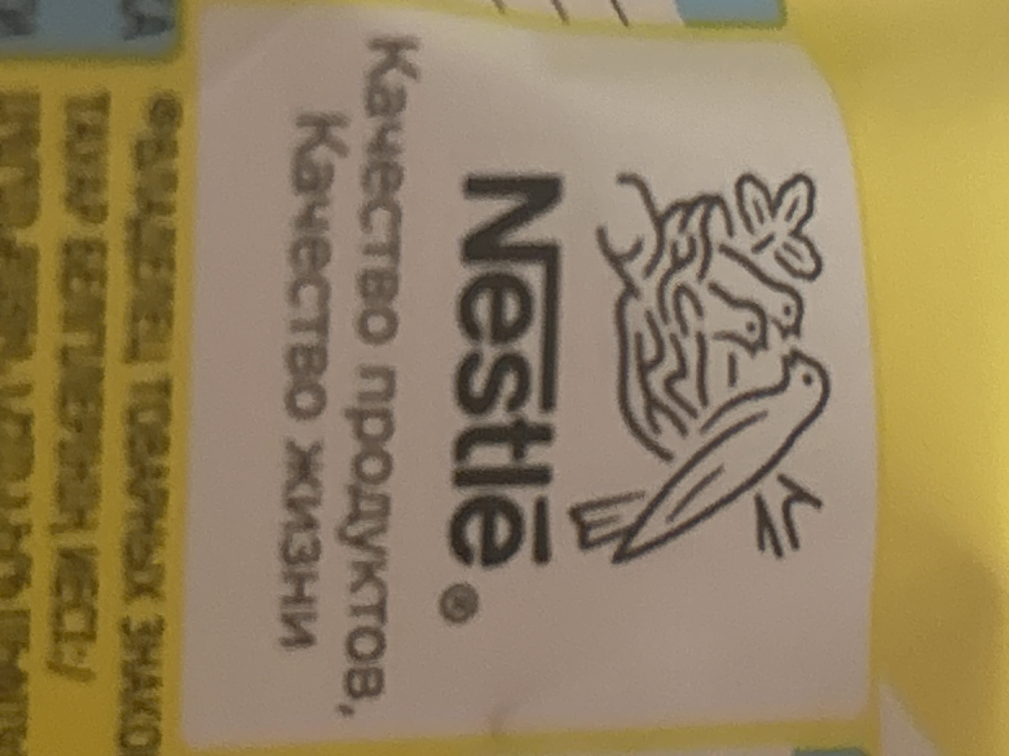 The image shows a close-up of a yellow package with a white label. The label features the Nestle logo, which is a bird's nest with birds and a bird on top. Below the logo, in Russian, it says "Качество продуктов, Качество жизни" which translates to "Quality of products, Quality of life". The bottom portion of the label has some text that is partially visible and appears to be in Russian as well. The image itself does not contain any information about the city of Minsk, Belarus.