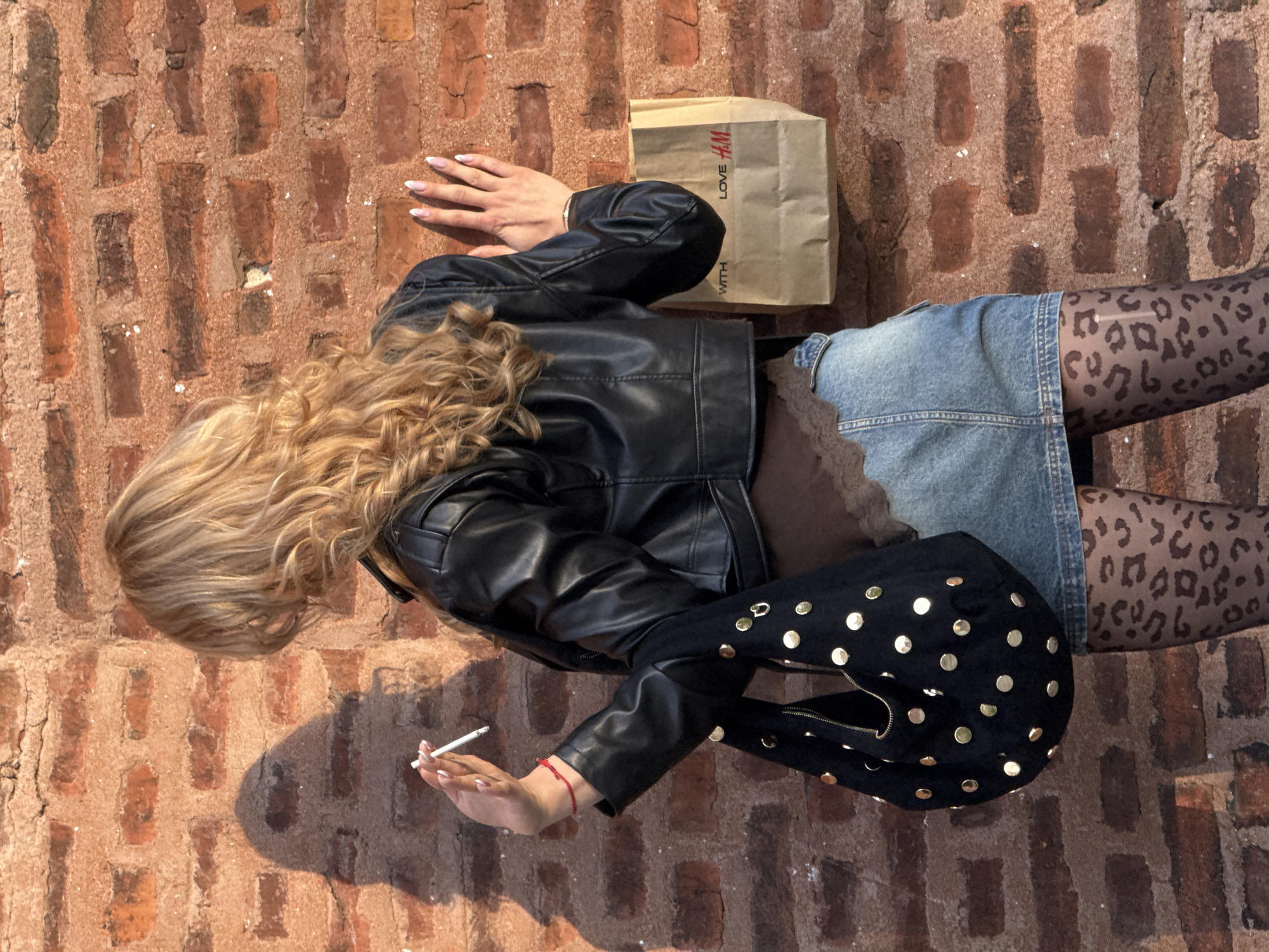 An overhead shot captures a person posing on a textured red brick surface under bright daylight, likely in Sofia, Bulgaria. The individual has long, wavy blonde hair and is dressed in a black faux leather jacket over a dark brown top with lace trim, a light blue denim mini-skirt, and sheer black tights with a leopard print pattern.
One hand, featuring long, light-tipped nails and a red string bracelet, holds a lit cigarette. Nearby, a brown paper shopping bag from H&M is visible, partially showing the text "LOVE H&M WITH." A black handbag adorned with numerous gold studs rests next to the person's leg. Strong sunlight casts distinct shadows, indicating a clear, sunny day. The overall scene suggests an urban outdoor setting.