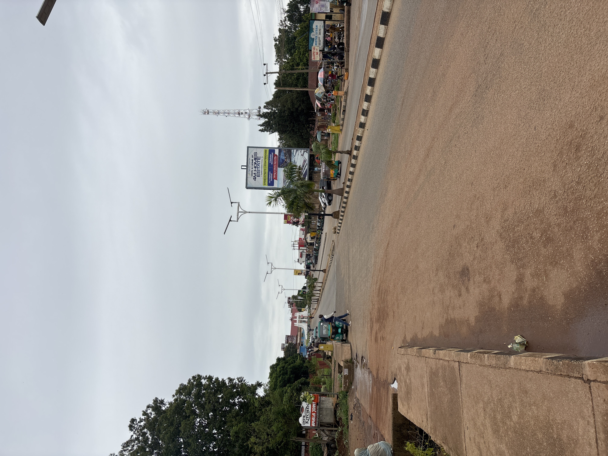 The image depicts a wide, paved road in Barakin Lerawi, Nigeria, under an overcast sky. The road is lined with a concrete curb and has a black and white striped border along its edge. On the left side of the road, a sidewalk area is visible, with a woman in a light blue garment sitting on a concrete structure. Adjacent to the sidewalk is a densely leaved tree. Further down the road, a green and white three-wheeled auto-rickshaw is parked, with a man in a blue shirt and jeans standing next to it, appearing to walk away.
On the right side of the road, there are several roadside stalls and shops. A tall communication tower is visible in the background. Various vehicles, including cars and more auto-rickshaws, are present in the scene. A billboard advertising "4M HOMES ESTATE" is prominently displayed. Palm trees are planted along the roadside. The overall scene suggests a moderately busy urban environment with commercial activity. Litter is visible on the ground near the curb in the foreground.