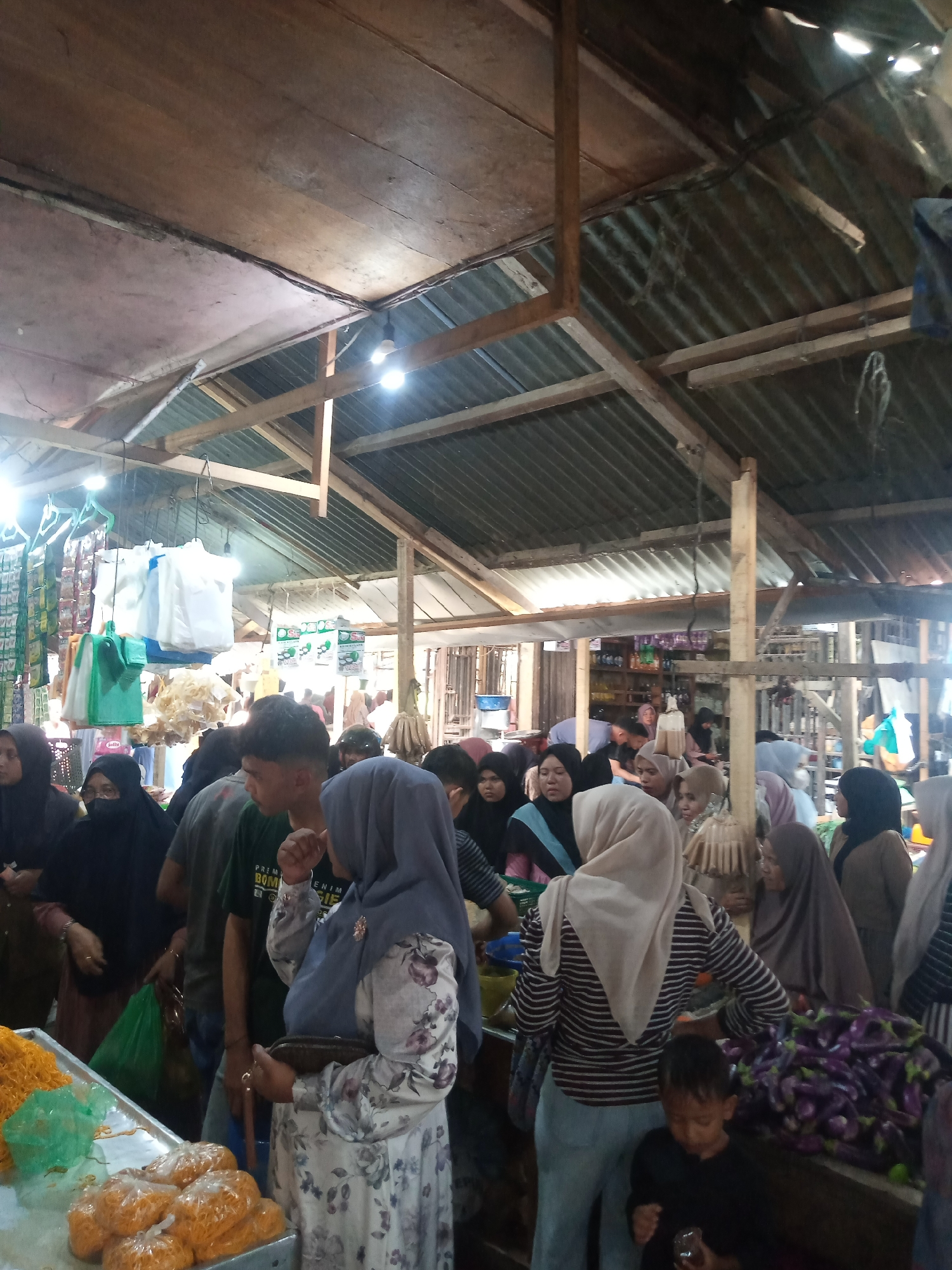 The image depicts a busy indoor market scene, likely in Grong-Grong, Indonesia. The market is covered by a corrugated metal roof supported by wooden beams and posts. Several bare light bulbs are suspended from the ceiling, providing illumination. The stalls are filled with various goods, including produce like eggplants piled high in the foreground. Packaged food items, plastic bags, and what appear to be dried noodles or snacks are also visible.
A crowd of people, predominantly women wearing hijabs in various colors, are browsing and interacting at the stalls. Some individuals are examining merchandise, while others are engaged in conversation. A young boy in a black shirt is visible in the lower right quadrant of the image, holding something small in his hand. The overall atmosphere suggests a typical bustling marketplace.
