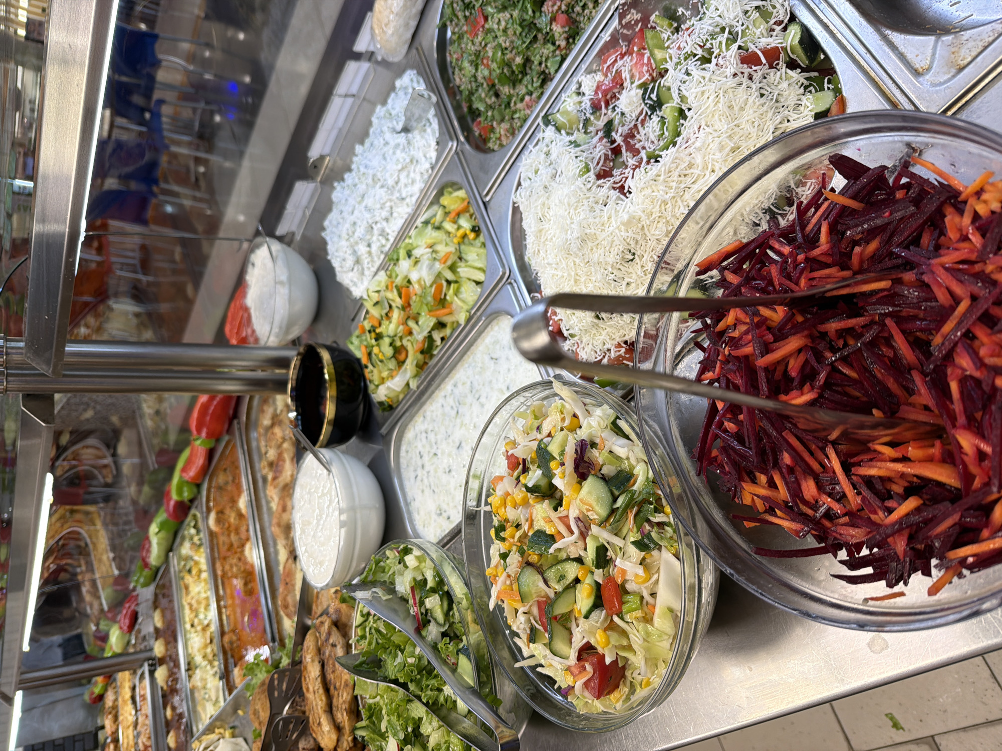 The image displays a close-up view of a buffet line, likely in a self-service restaurant. Several stainless steel pans are filled with various food items. In the foreground, two glass bowls are prominent. The left bowl contains a salad of shredded carrots and beets, with tongs resting inside. The right bowl holds a mixed salad with chopped cucumbers, tomatoes, corn, and what appears to be cabbage. Behind these, a larger stainless steel pan is filled with shredded white cheese over a bed of chopped vegetables including tomatoes and cucumbers. To the left of the cheese, a pan holds a salad of chopped lettuce, cucumbers, and radishes, with tongs. Further back, a pan contains a creamy white salad with visible herbs, possibly a tzatziki or yogurt-based salad. Next to that, a pan is filled with a vegetable medley of chopped zucchini, carrots, and corn. To the right, a pan holds what looks like a grain salad with chopped parsley and tomatoes. In the background, more food pans are visible, including what appear to be cooked meat dishes. The overall environment suggests a food service establishment with a focus on fresh salads and prepared meals. This scene is located in the food court area of the Mall of Plovdiv in Plovdiv, Bulgaria. There are no people clearly visible in the image.