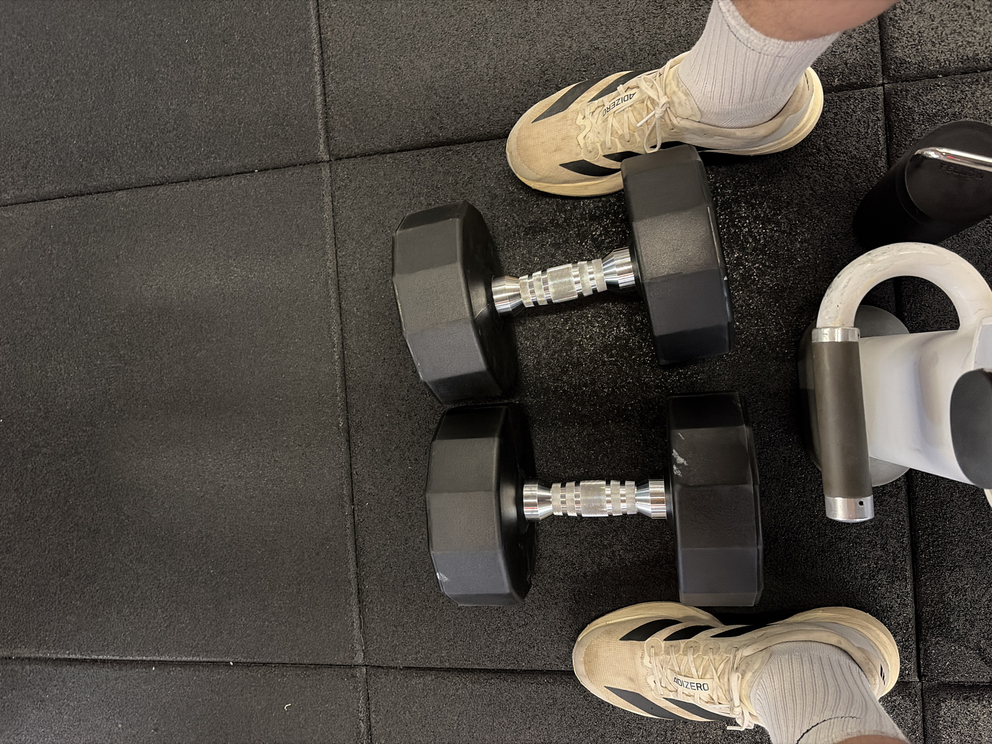 The image shows a section of a gym floor in what appears to be New York City, United States of America, with black rubber tiling. Two black dumbbells, each with a textured silver handle, are lying on the floor. A pair of athletic shoes, marked "ADIZERO," are visible in the top right and bottom right corners of the image, each appearing to be worn by someone standing near the dumbbells. The person is wearing white athletic socks. Parts of a weightlifting apparatus, including a white curved handle and a grey vertical support, are visible in the right corner.
