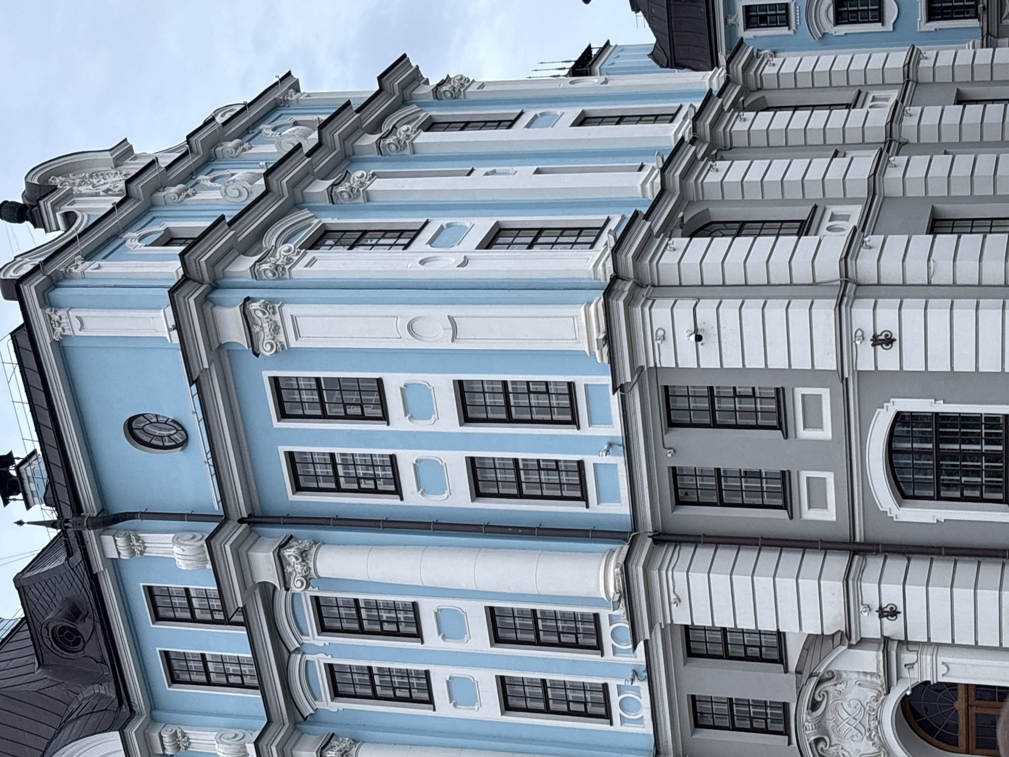 This is a medium-angle, exterior, eye-level, vertical shot of the Baroque-style architecture of the Smolny Cathedral in Saint Petersburg, Russia. The light blue and white facade of the building is the dominant subject, featuring rows of rectangular windows with dark frames and a decorative cornice. The sky is overcast, suggesting a cloudy day. The building's ornate details, including pilasters, curved pediments, and carved ornamentation, are visible. The perspective is from a low angle, emphasizing the height and grandeur of the structure. No people or activities are depicted. The image is a static architectural study, focusing on the building's design and materials. The time of day is indeterminate due to the overcast conditions. There is no visible text.